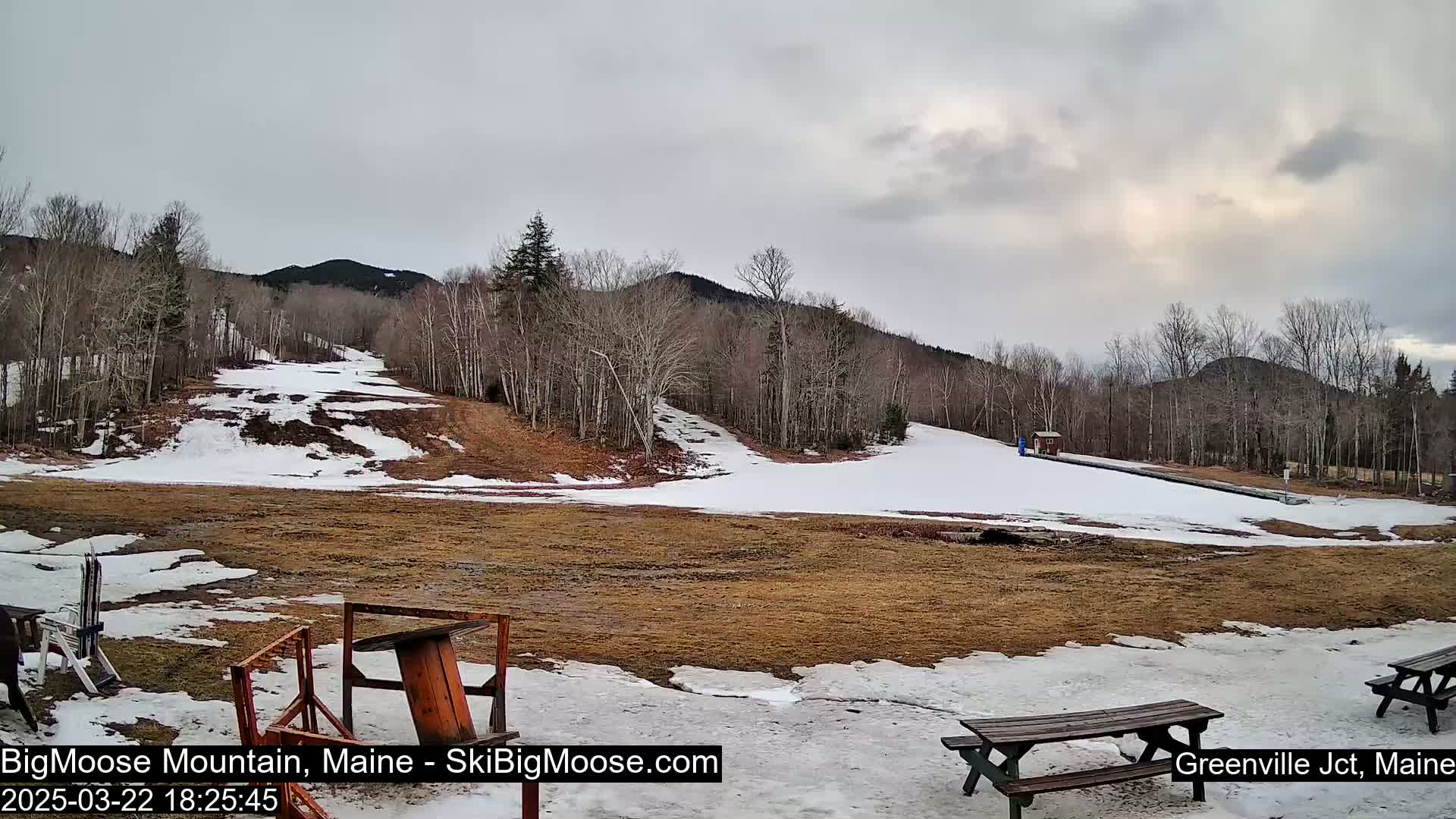 A mostly cloudy day reveals a snow-covered ski slope at the base of a mountain, with patches of brown grass and melting snow visible at the bottom.
