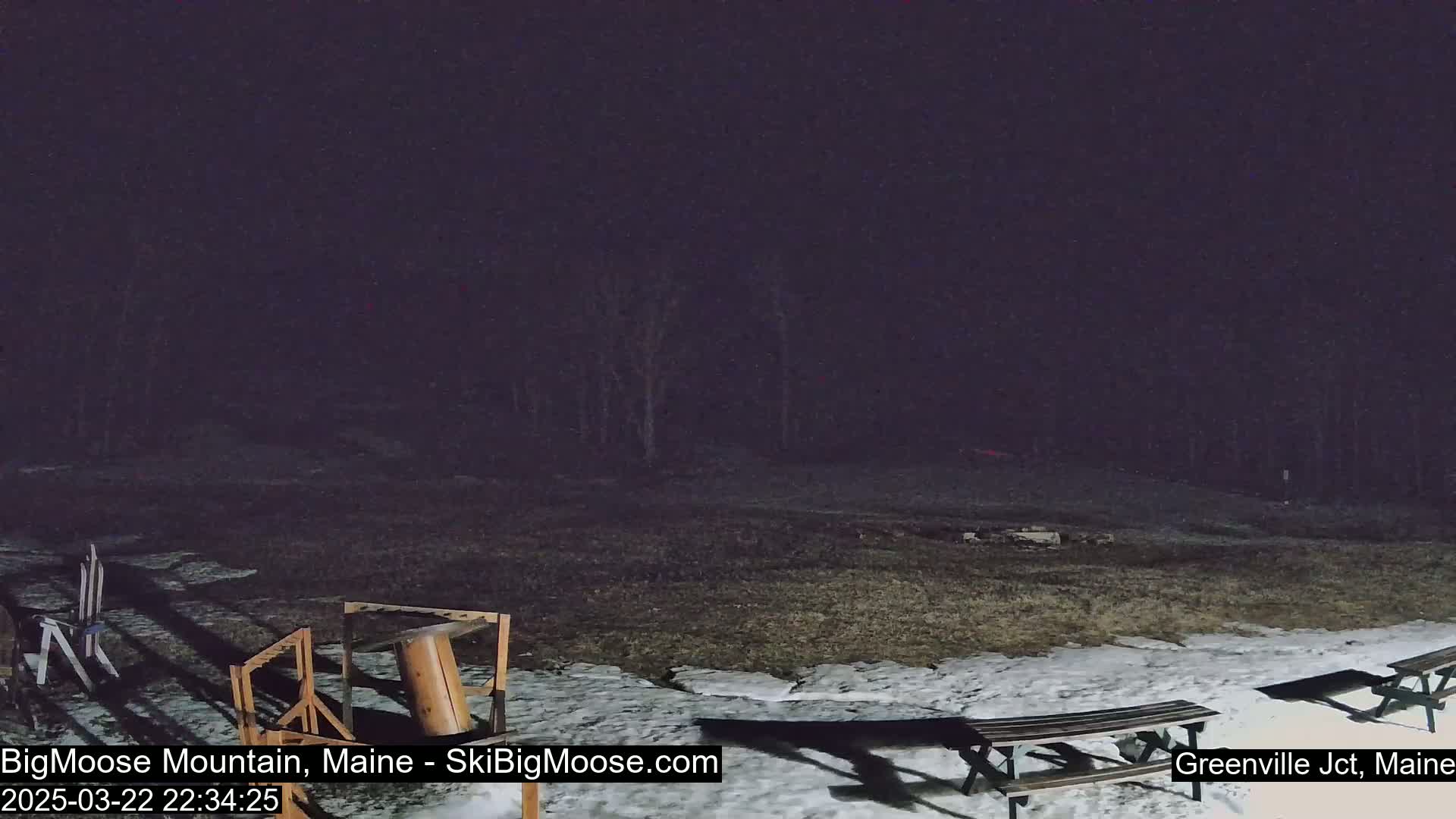 A nighttime scene shows a mostly snow-covered grassy area with picnic tables, some wooden structures, and trees in the background under a dark sky.