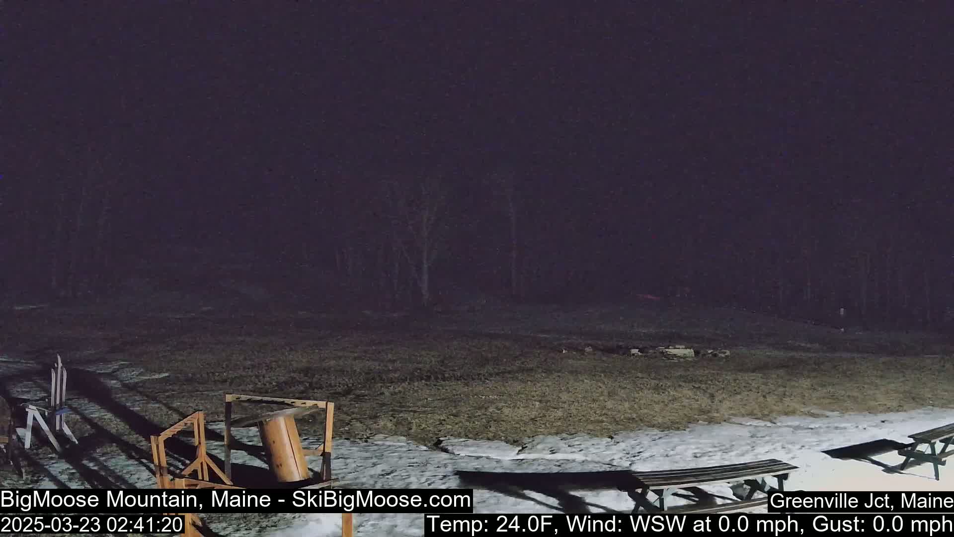 A nighttime outdoor scene shows a mostly barren, snow-dusted field with some wooden structures and picnic tables, under a dark sky with calm weather.