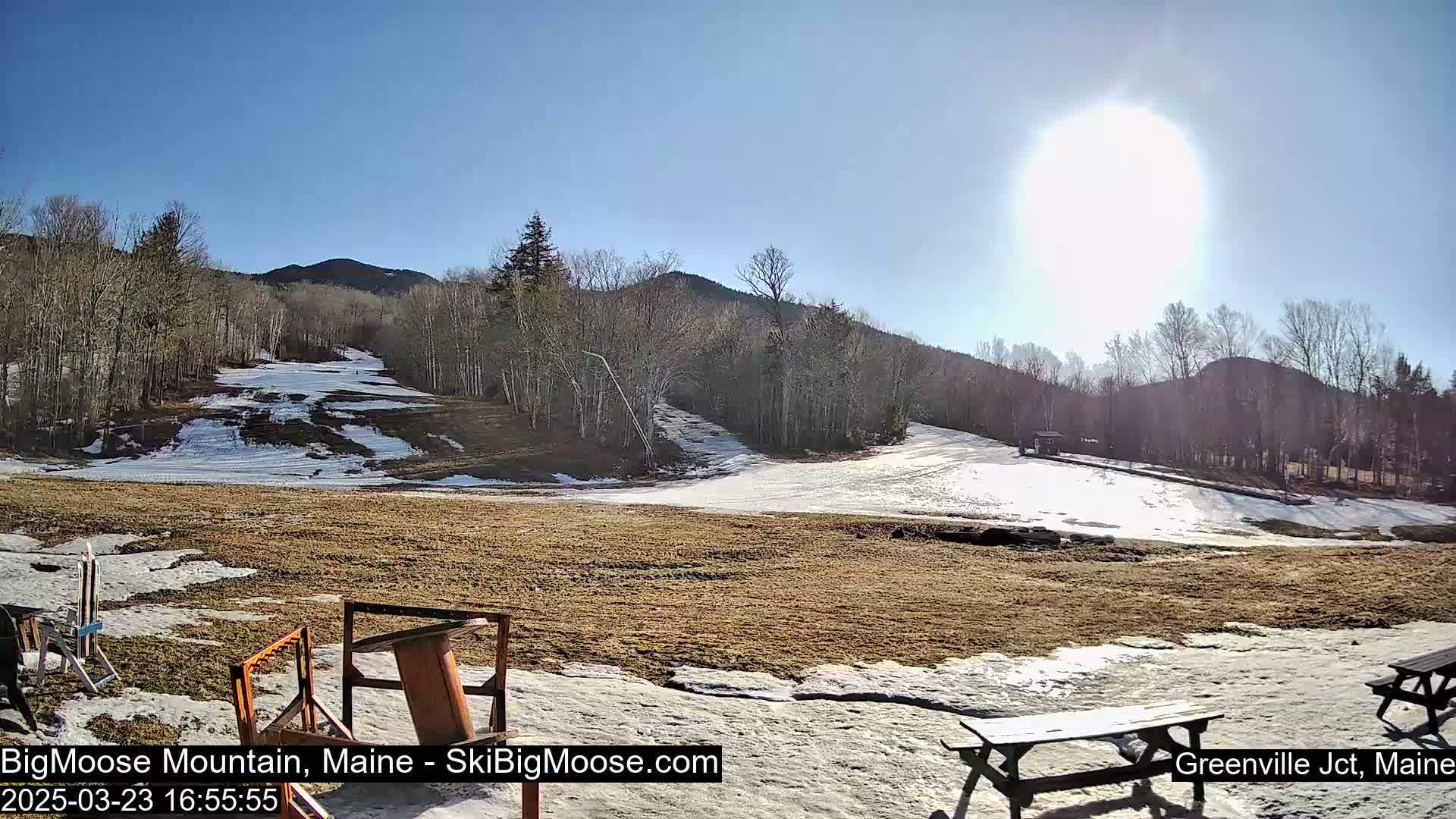 A sunny day reveals a partially snow-covered ski slope nestled among bare trees and hills.