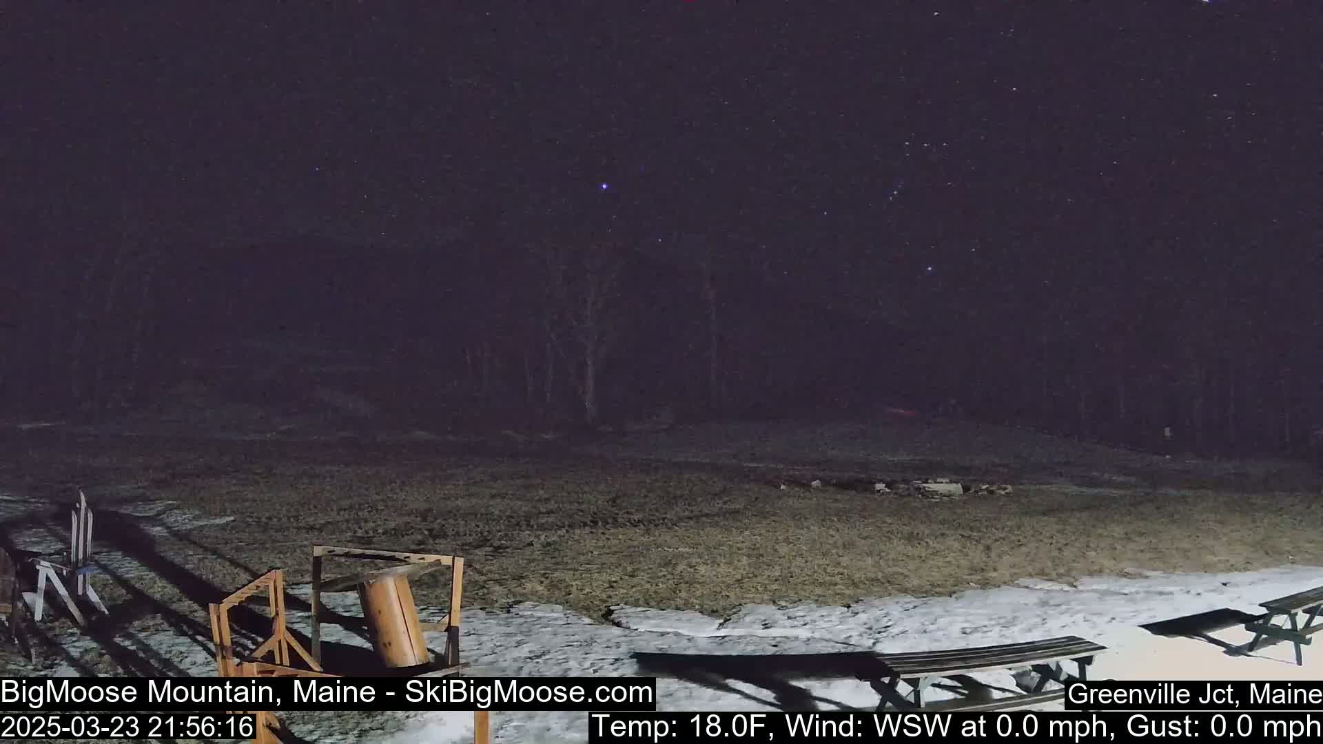 A nighttime outdoor scene shows a mostly clear sky with stars, a sparsely snow-covered ground with picnic tables, and some wooden structures.