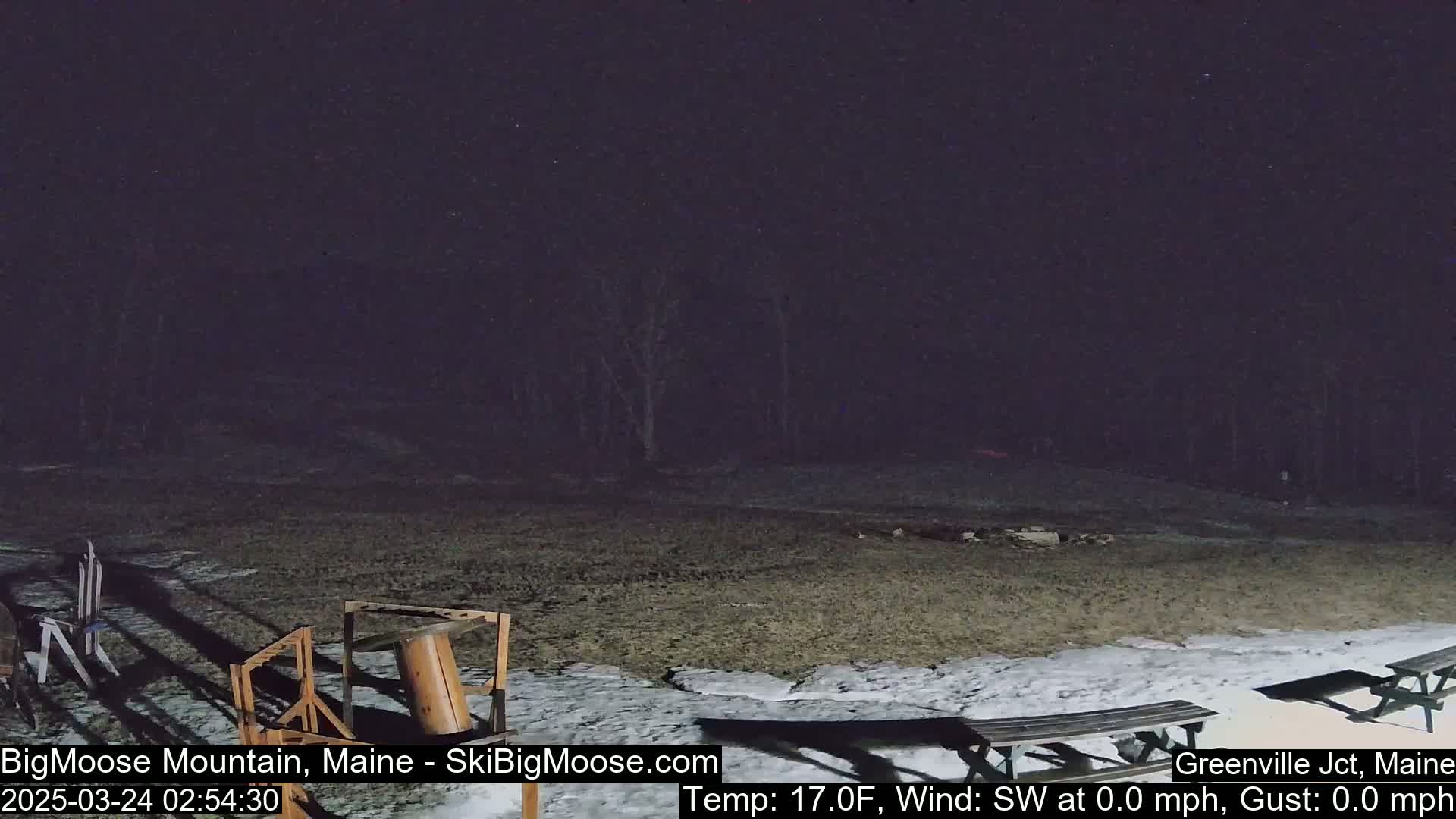 A nighttime outdoor scene shows a mostly bare, snow-dusted ground with some wooden picnic tables and equipment, under a dark, clear sky.