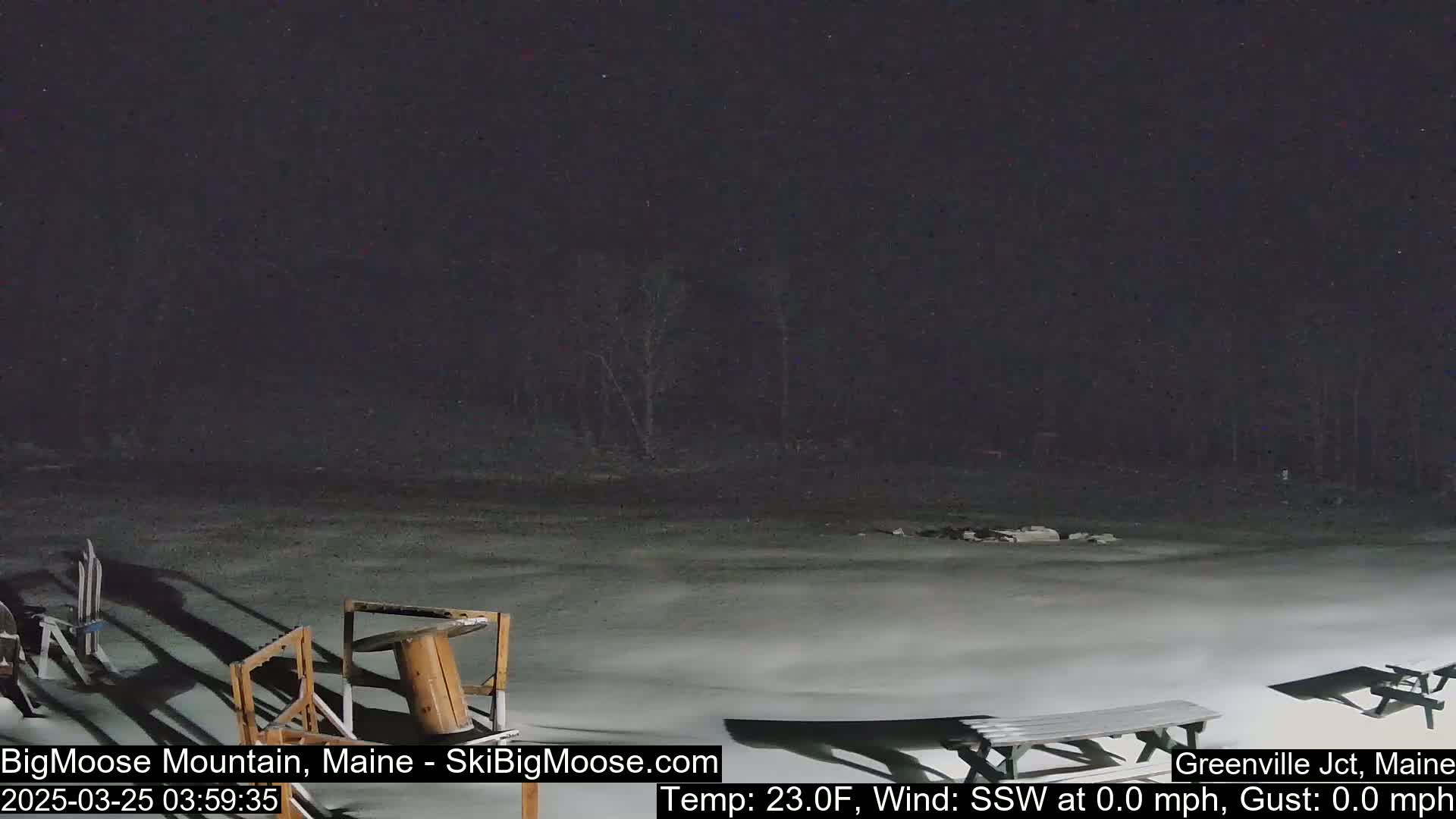 A nighttime, snow-covered outdoor scene shows a mostly empty paved area with some picnic tables and other outdoor furniture, under a dark sky with a calm wind.