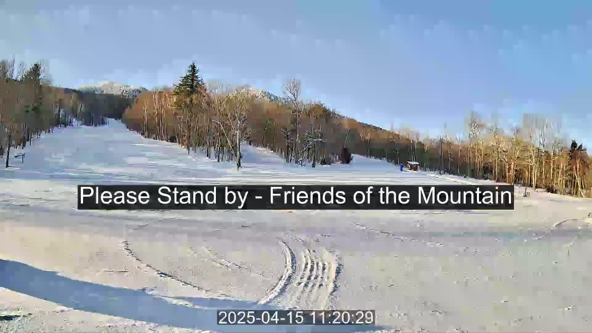 A snow-covered landscape shows a mostly clear, sunny day with a view of a mountain in the background and leafless trees along the sides.
