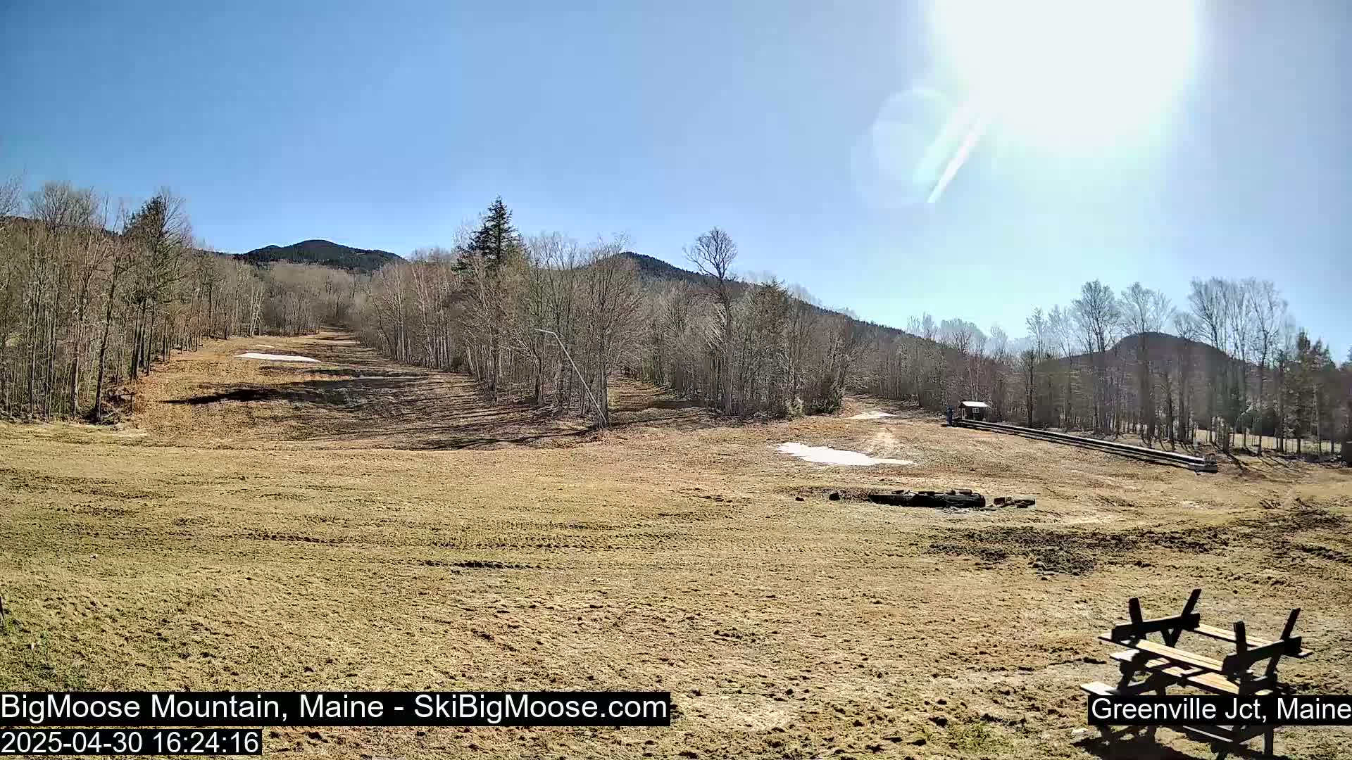 A mostly sunny day reveals a barren ski slope with a picnic table in the foreground, surrounded by leafless trees and distant mountains.