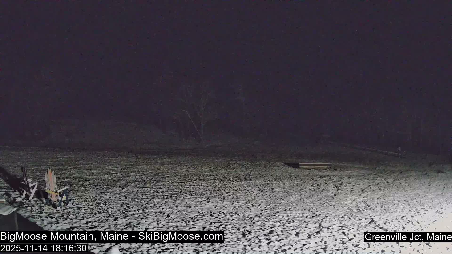 The dark outdoor scene at night shows a snow-covered ground with several chairs on the left and a bare tree visible in the distance.