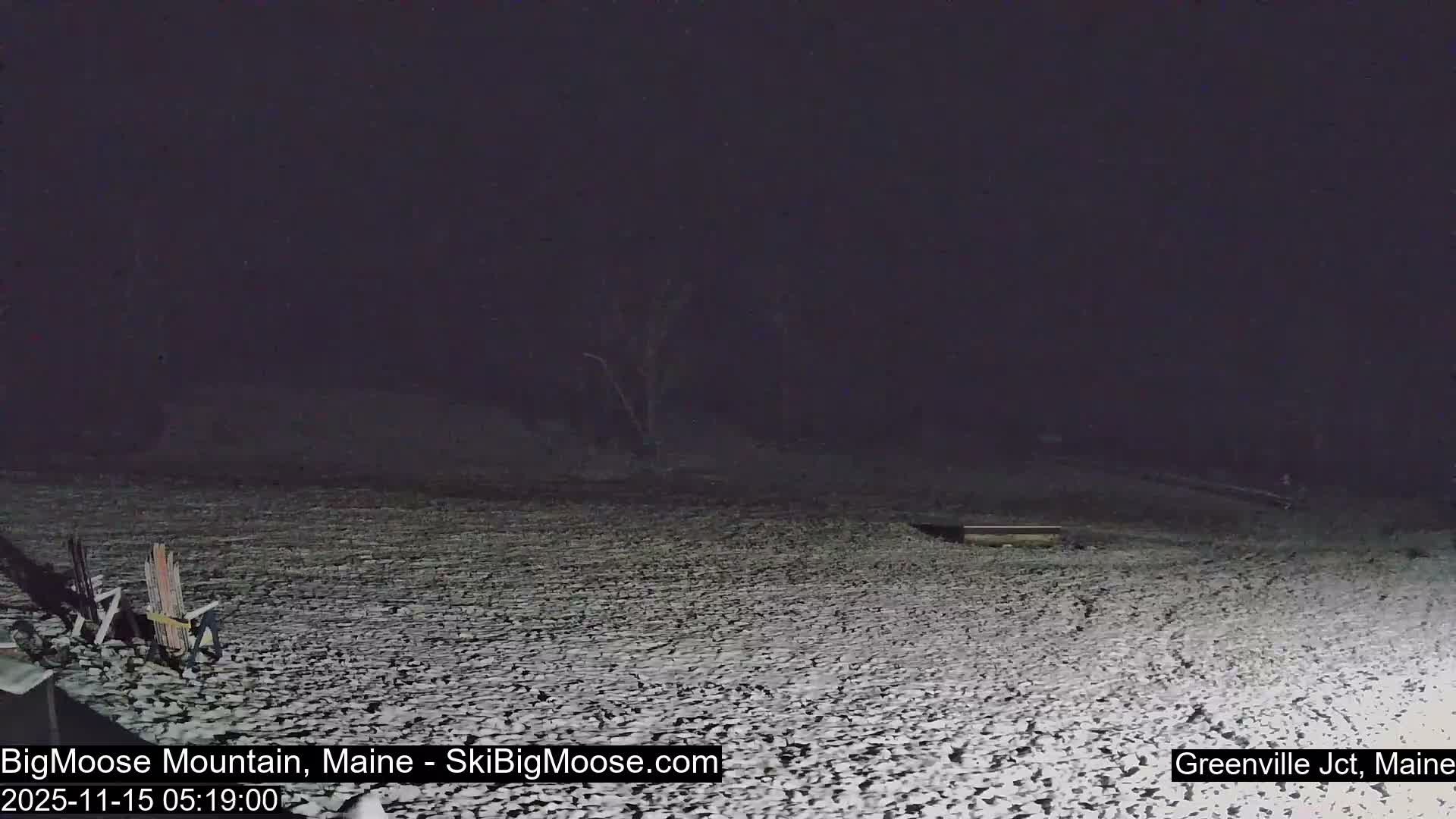A dark winter night scene reveals a snow-covered ground with bare trees in the background, faintly illuminated by an unseen light source that casts shadows from two chairs on the left, under clear or overcast skies.