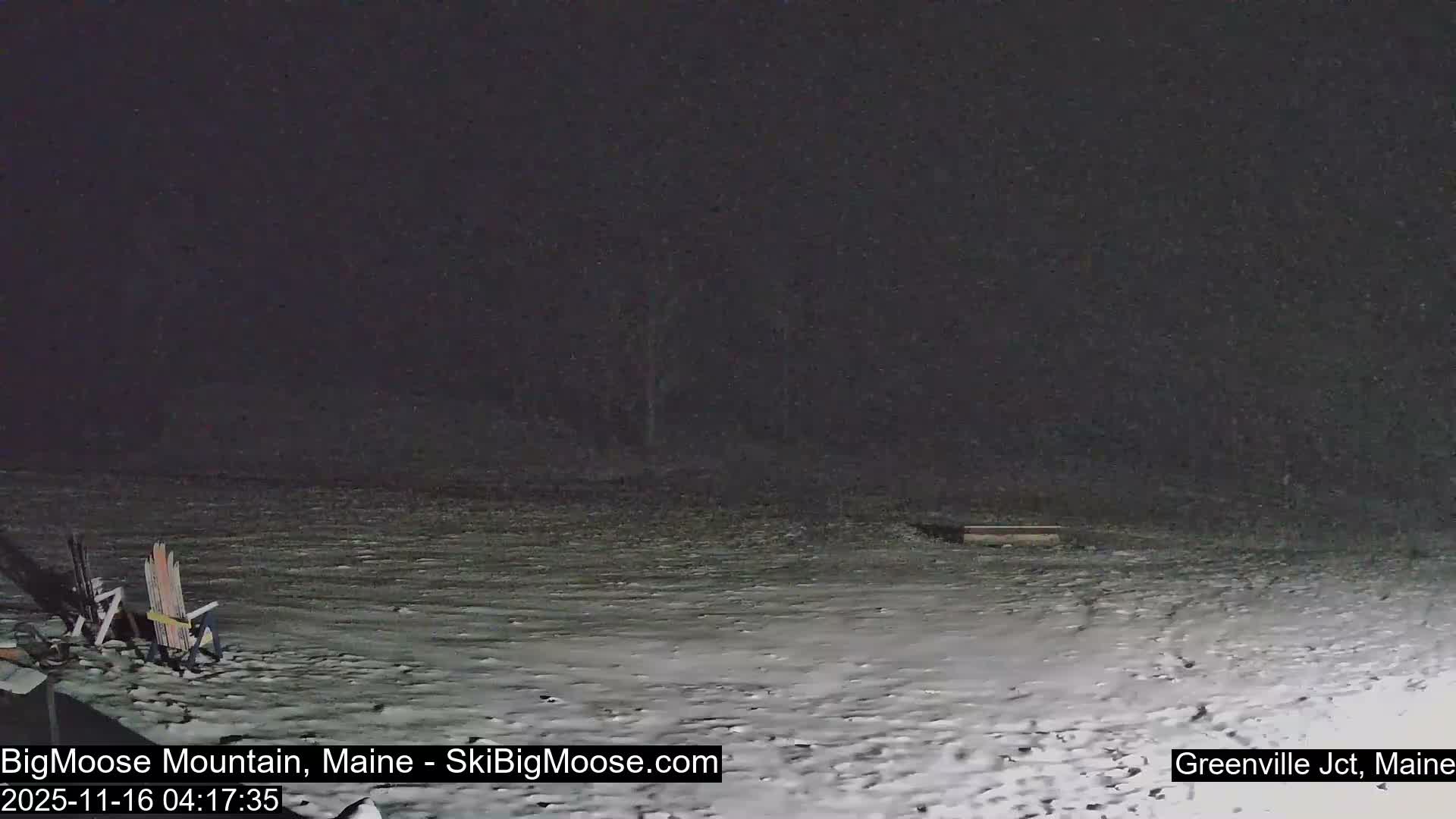 Two Adirondack chairs sit on snow-dusted ground in a dark outdoor scene under clear night skies.