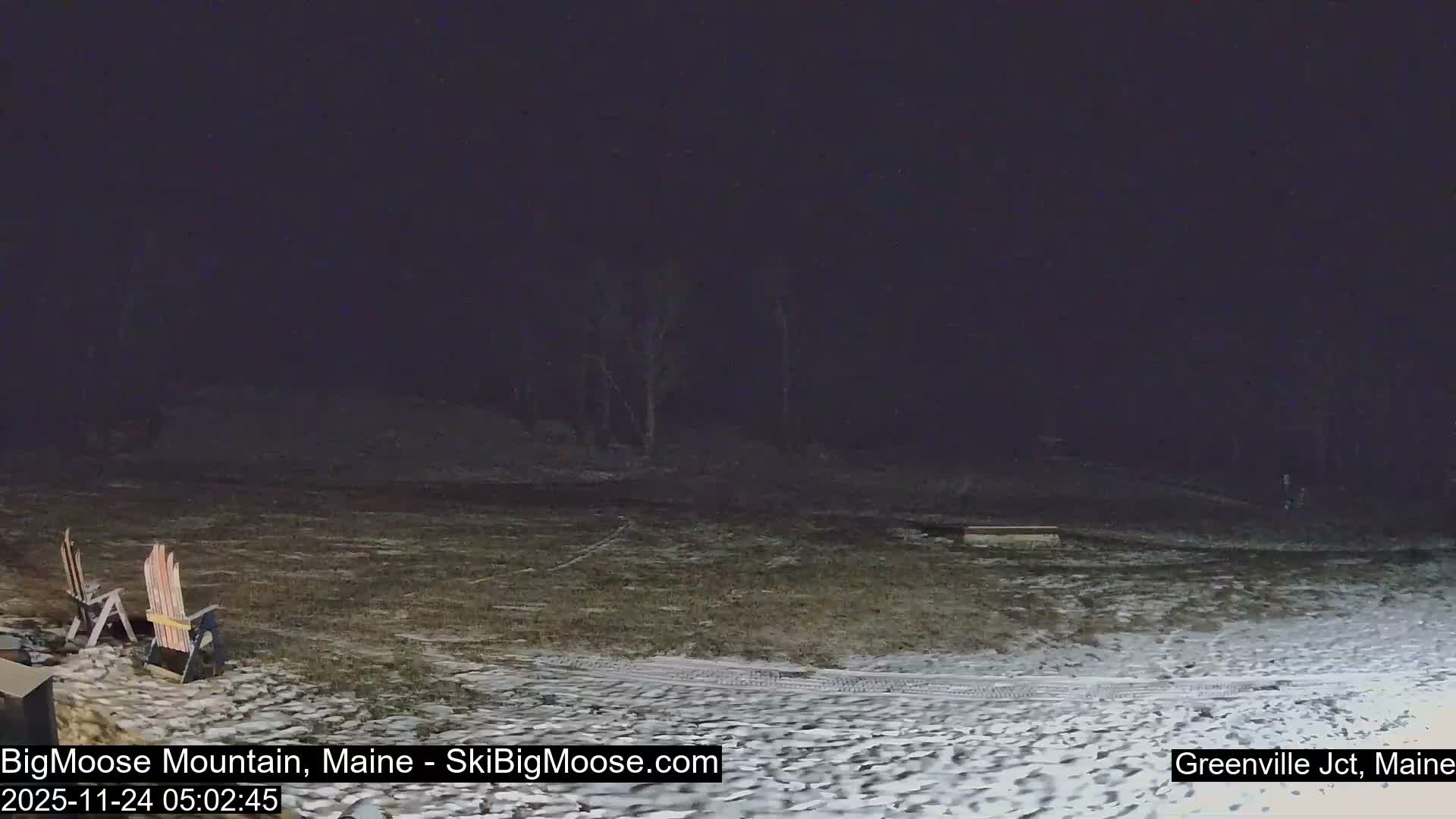An outdoor scene on a cold, clear night shows patchy snow covering the ground with visible tire tracks, bare trees in the background, and two Adirondack chairs to the left.