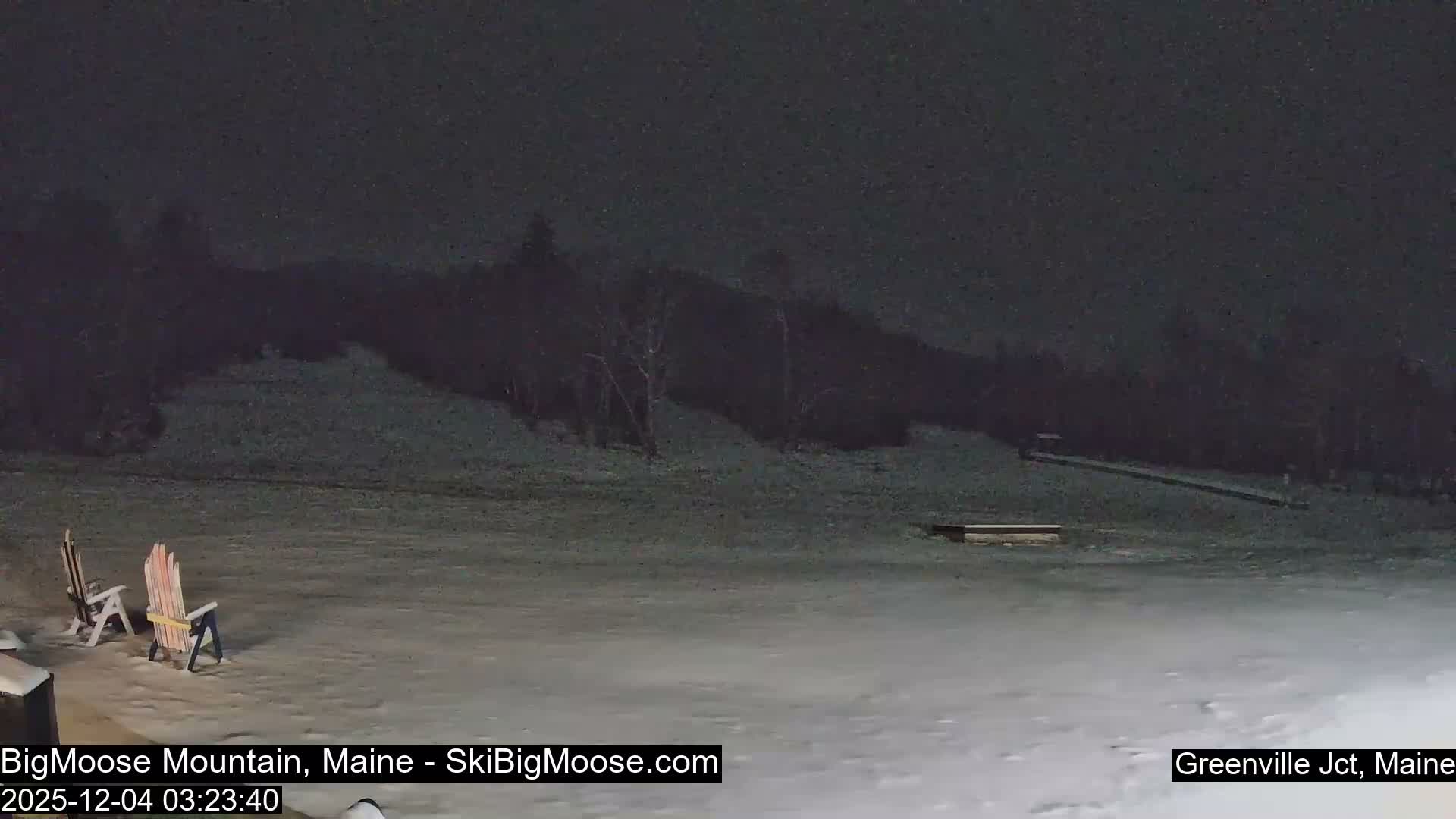 The image depicts a snow-covered outdoor scene at night, featuring two Adirondack chairs in the foreground and a dark, forested hillside in the background under clear skies.