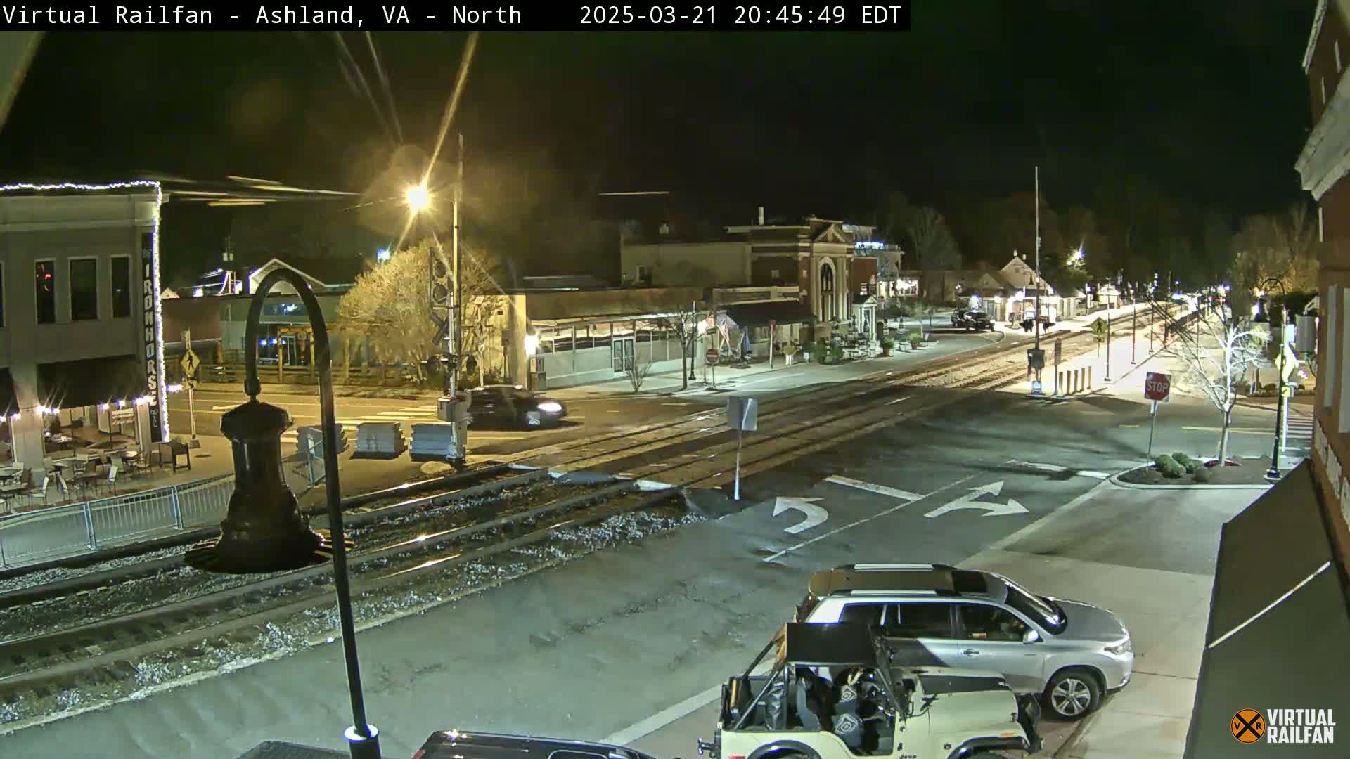 A nighttime street scene shows a railroad crossing near a town square with several cars parked and driving, under clear skies.