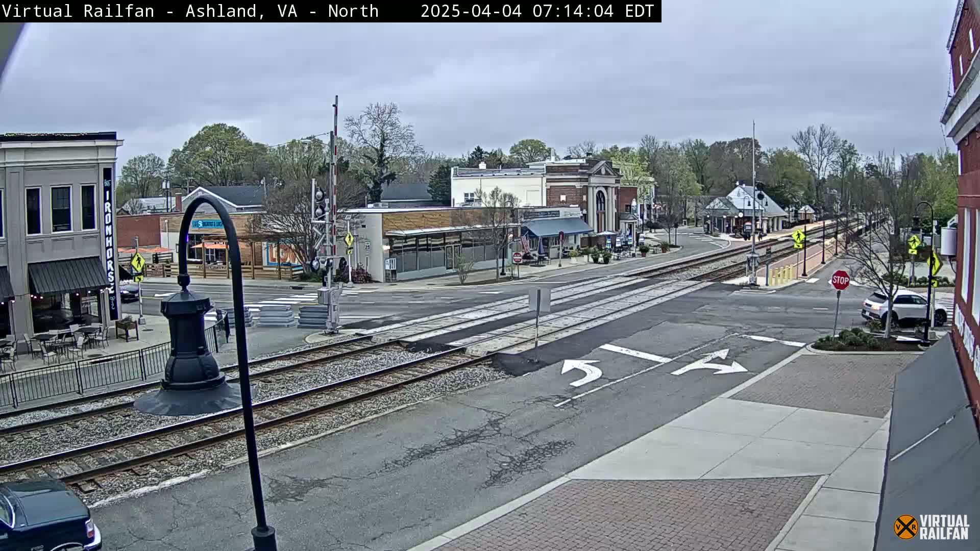 A street scene on an overcast day shows a railroad crossing in front of several small buildings, with cars parked along the street and one car stopped at the crossing.