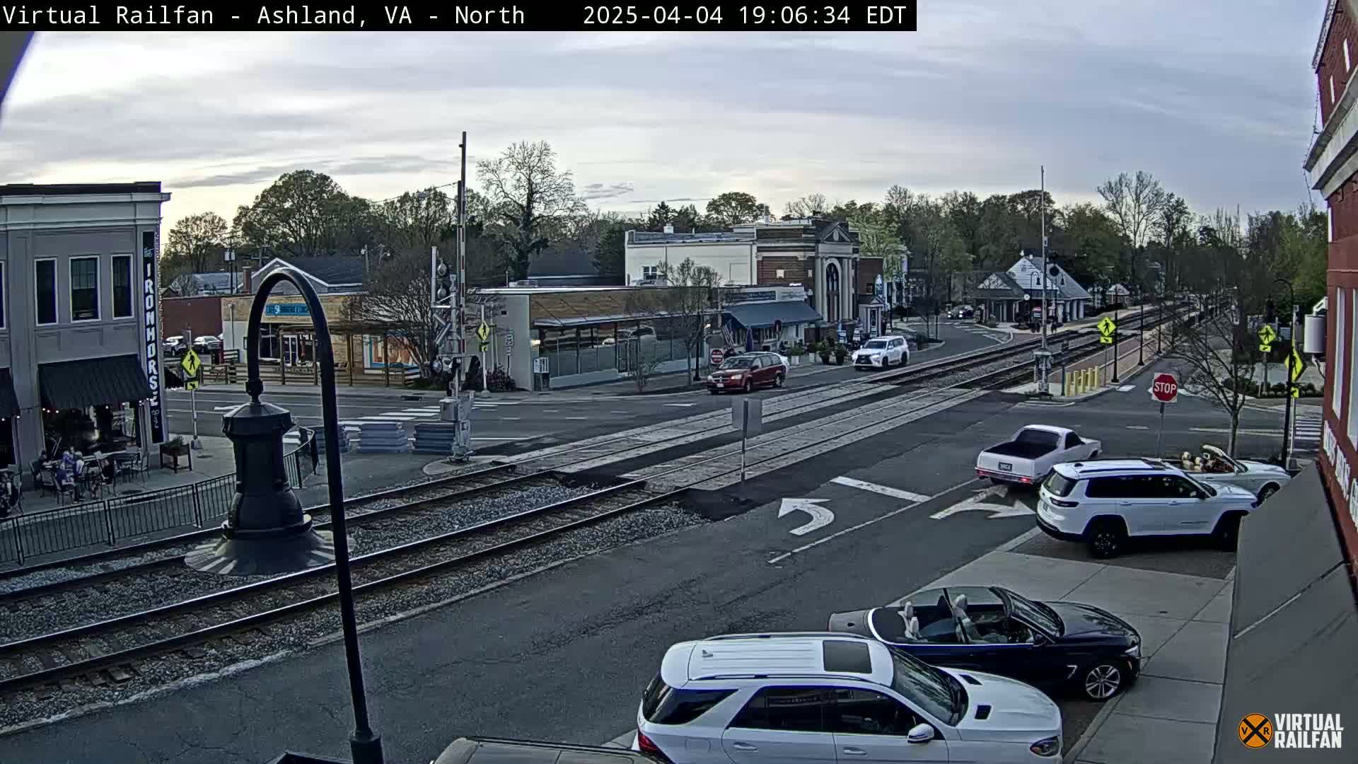 A street scene in a town, under an overcast sky, shows several vehicles at an intersection near railroad tracks.