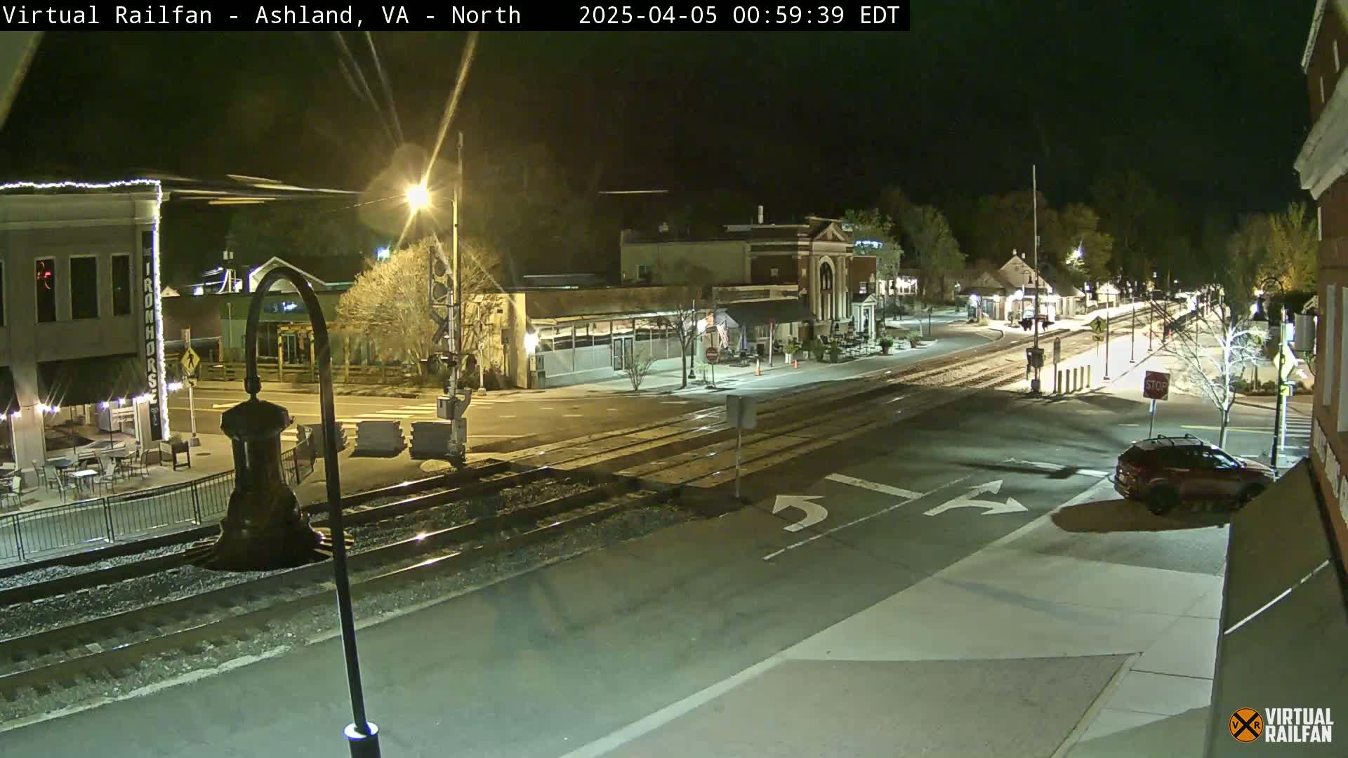 A nighttime view of a town's street intersection with train tracks, showing buildings, streetlights, and a vehicle.
