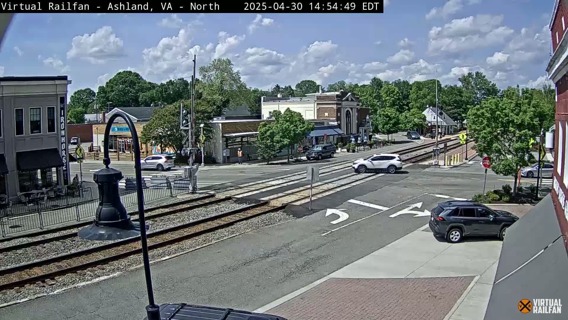 A partly sunny day shows a street intersection with railroad tracks, several cars, and buildings in a small town.