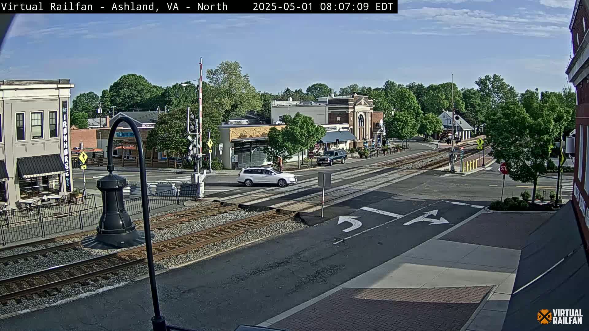 A sunny day shows a street intersection with railroad tracks, several buildings, and a few cars, one of which is white.