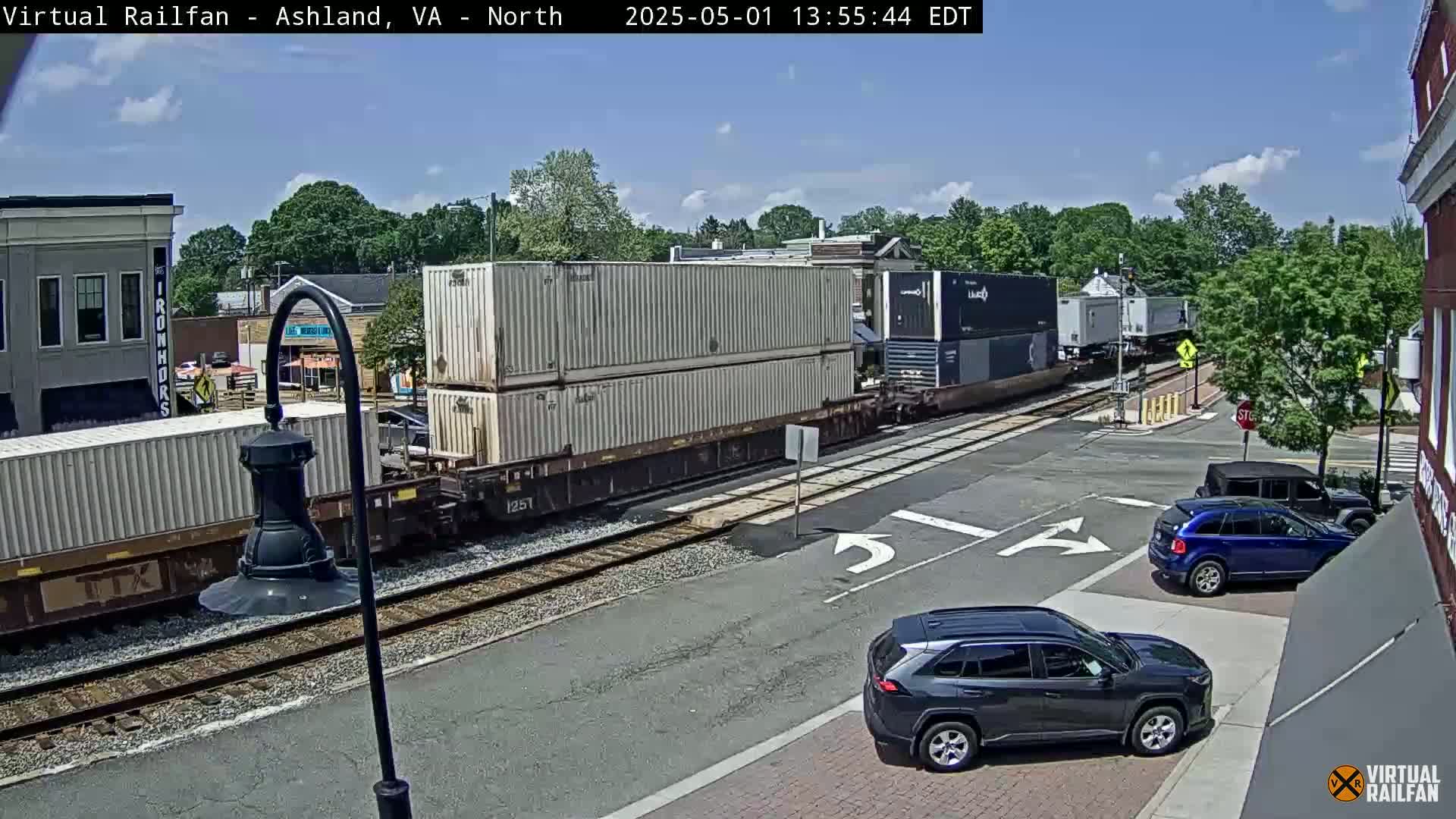 Under a partly cloudy sky, a freight train carrying multiple shipping containers passes through a street intersection where several cars are stopped.