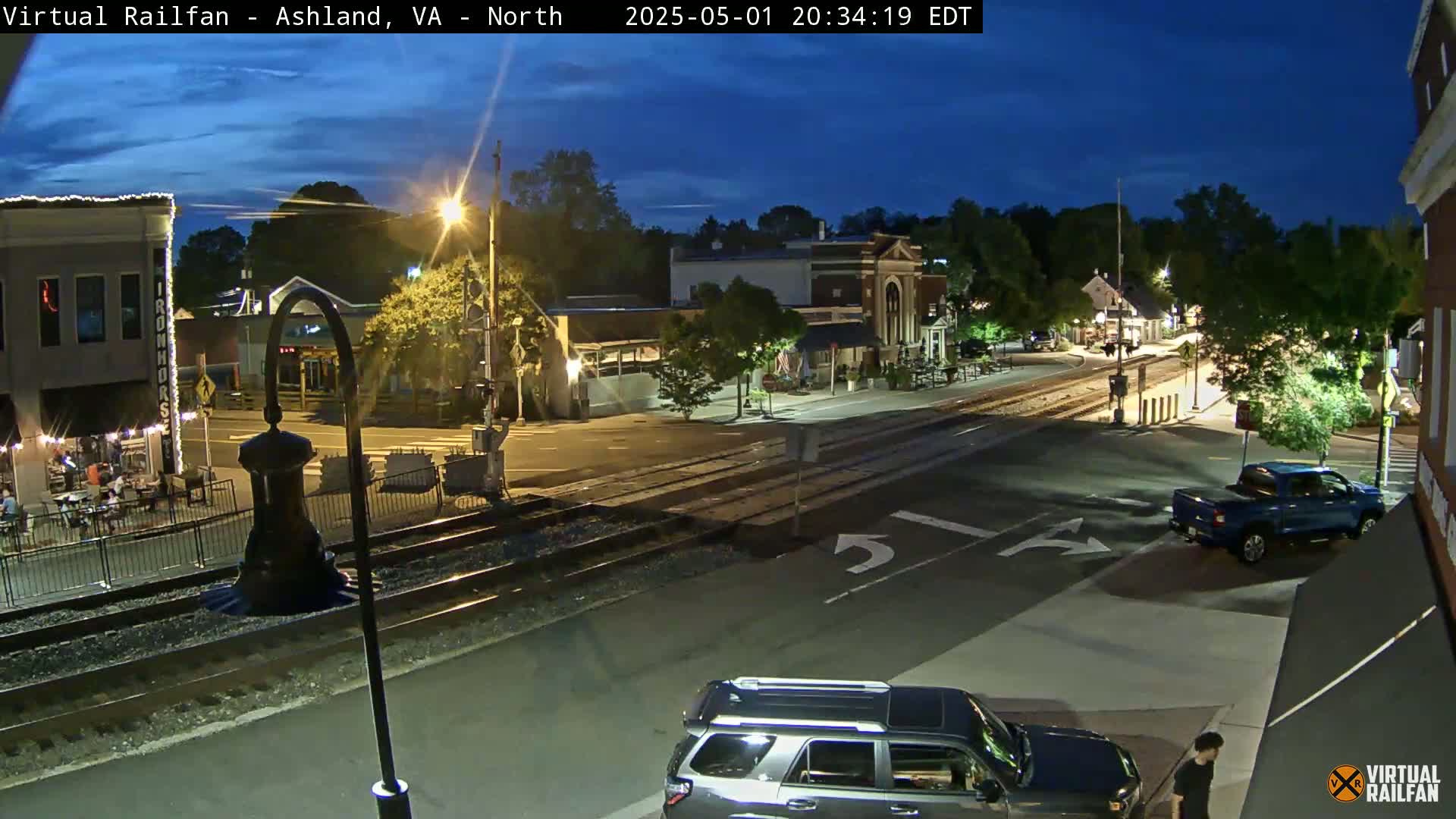 A nighttime view of a town's intersection with railroad tracks, featuring several buildings, vehicles, and a few pedestrians under a mostly clear twilight sky.
