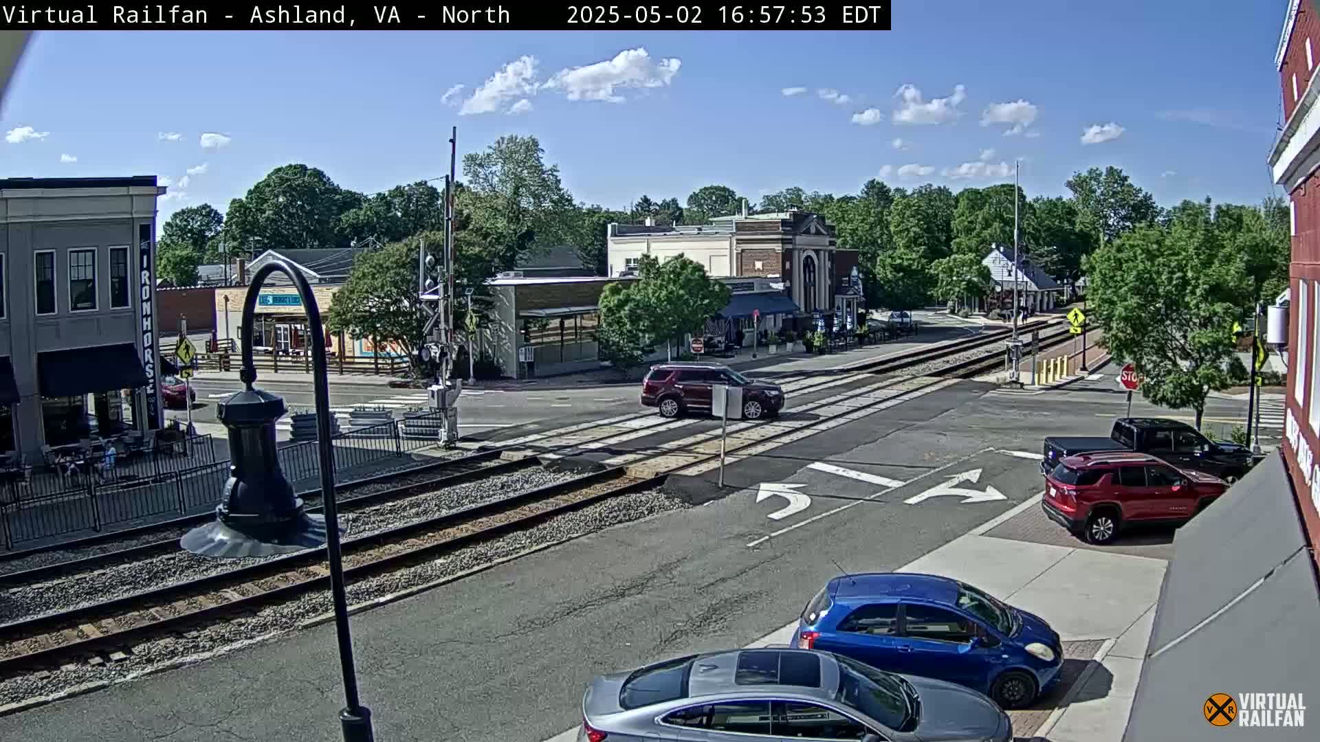 A sunny day view of a town square with railroad tracks crossing a street, several cars, and buildings including shops and restaurants.