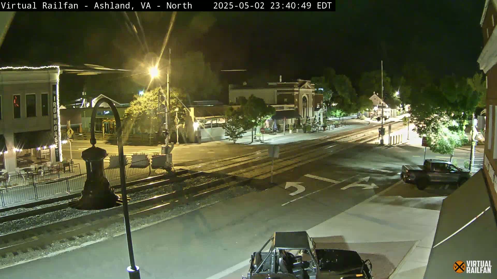 A nighttime view of a town's street intersection with train tracks, buildings, and a few vehicles present.