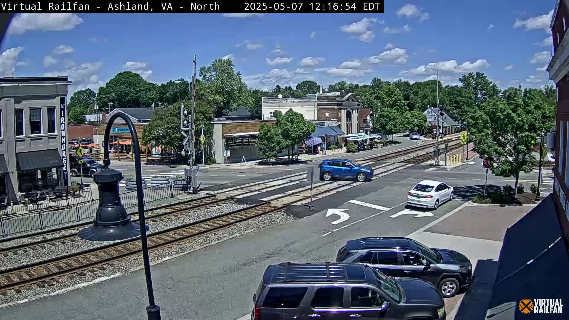 A sunny day shows a street scene with several cars at an intersection near train tracks, with buildings and trees in the background.