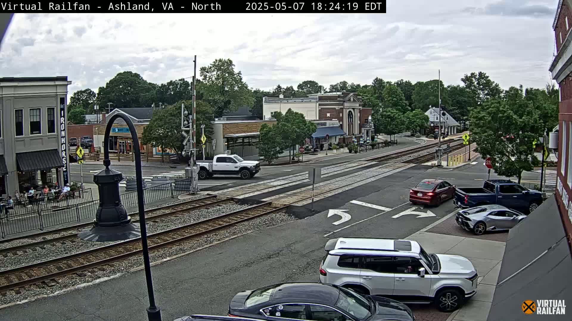 A mostly cloudy day reveals a town square with railroad tracks intersecting a road, several cars, and pedestrians enjoying outdoor seating at nearby businesses.