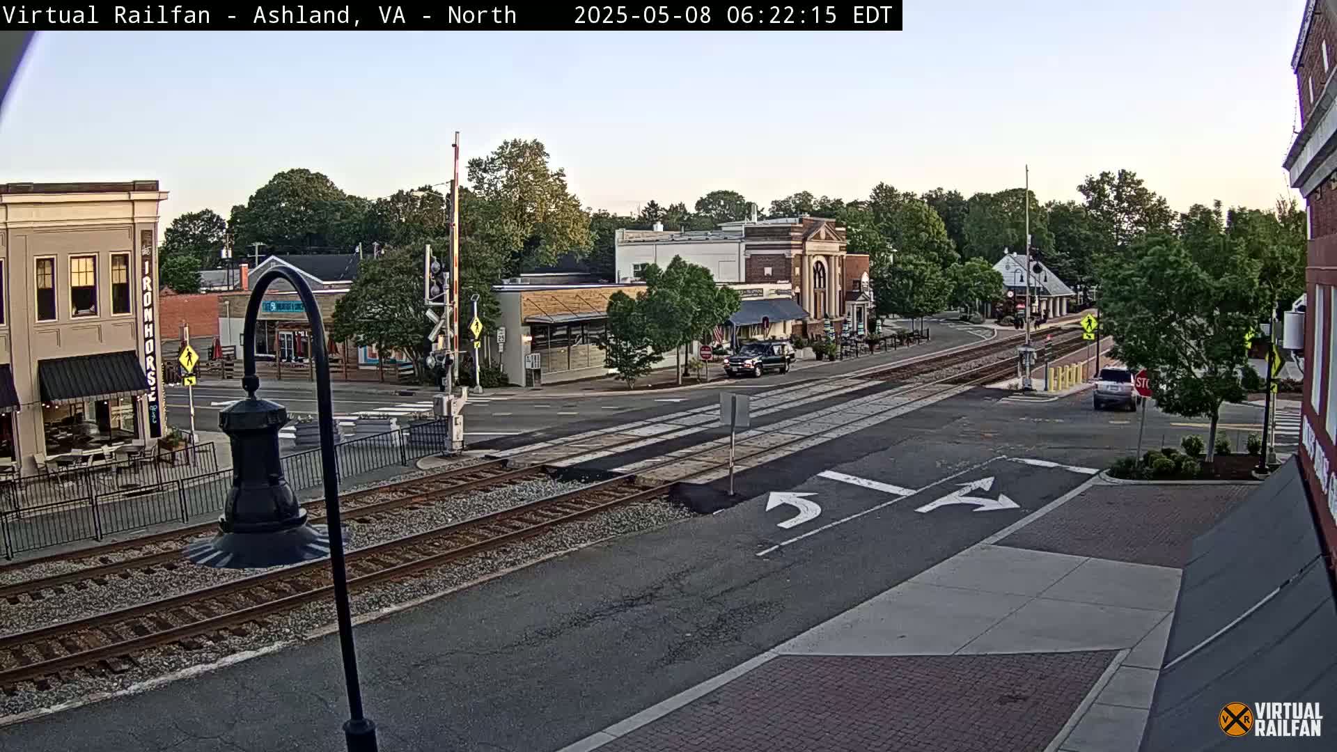A street-level view of a town's railroad crossing intersection on a partly cloudy morning, with several cars visible and businesses lining the street.
