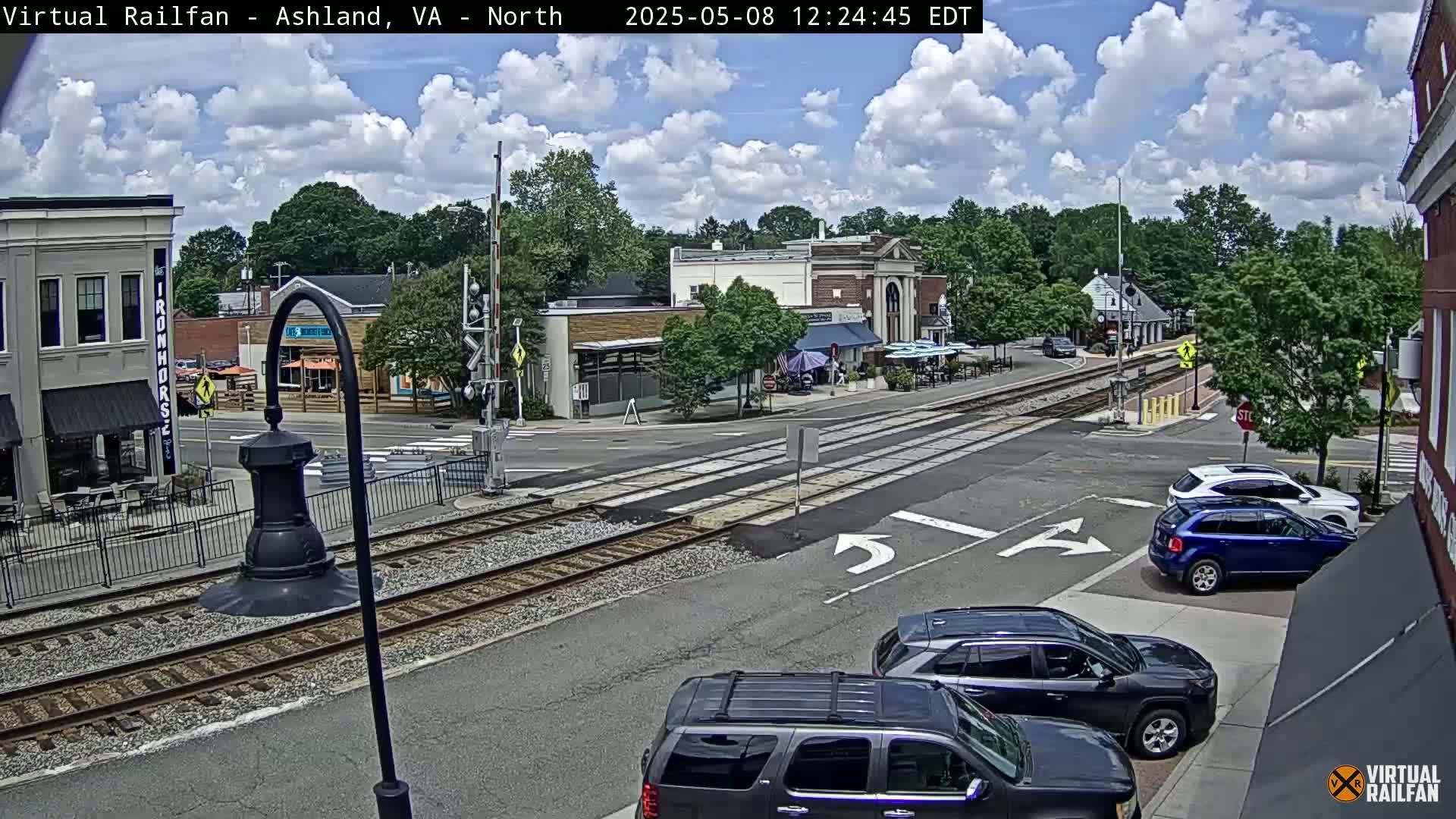 A partly sunny day reveals a street intersection with railroad tracks, several cars, and a collection of buildings including shops and restaurants.