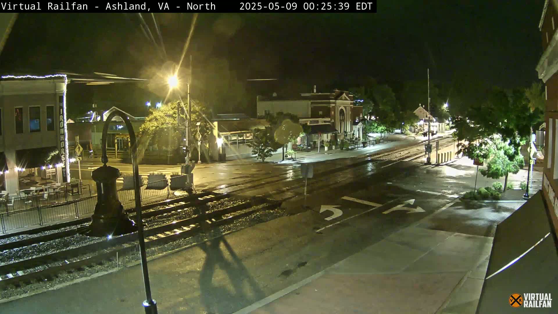 A nighttime view of a wet street intersection with train tracks, streetlights, and buildings along the sides.