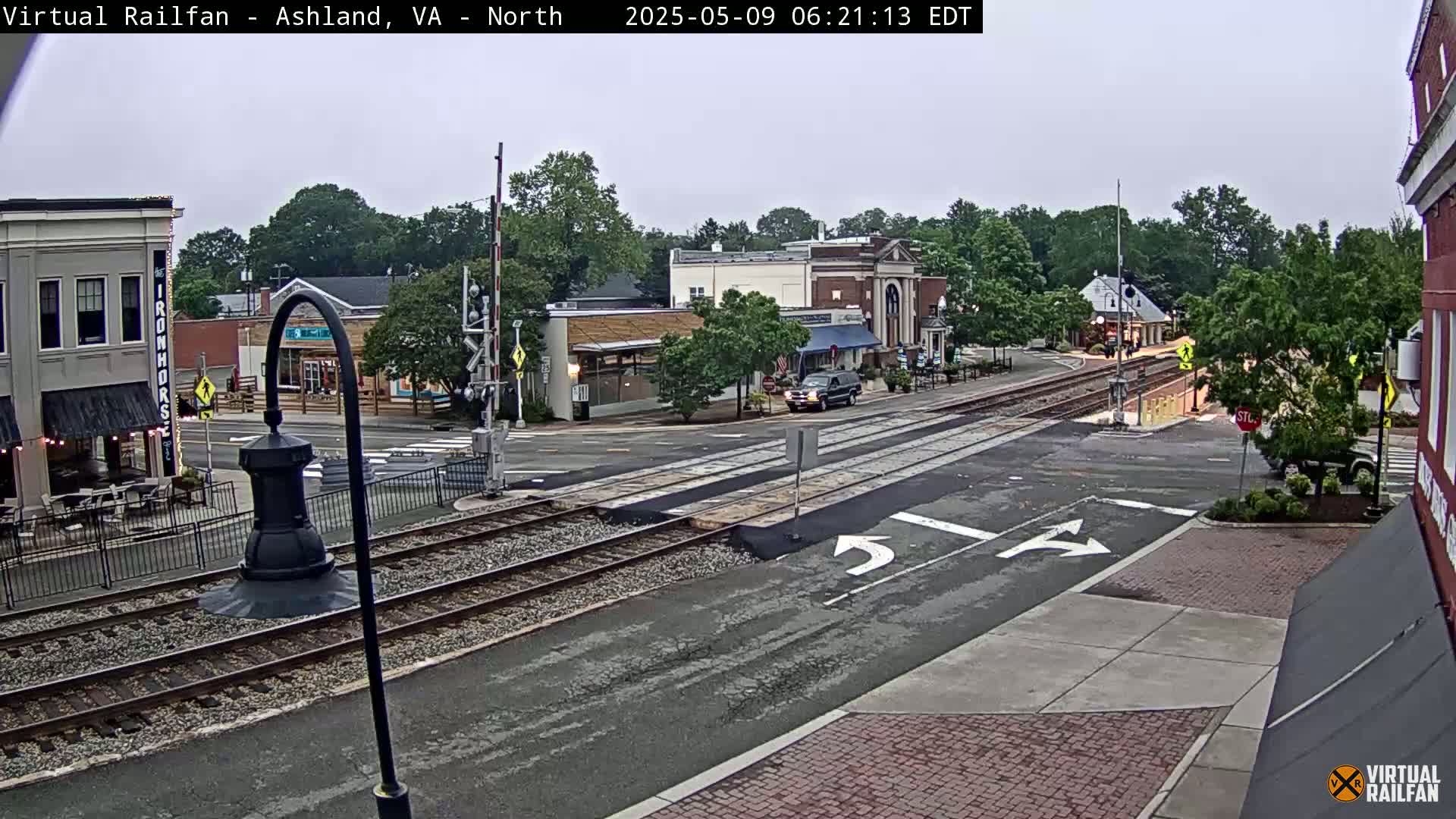 A mostly cloudy daytime scene shows a railroad crossing in a small town with a few cars and buildings visible.