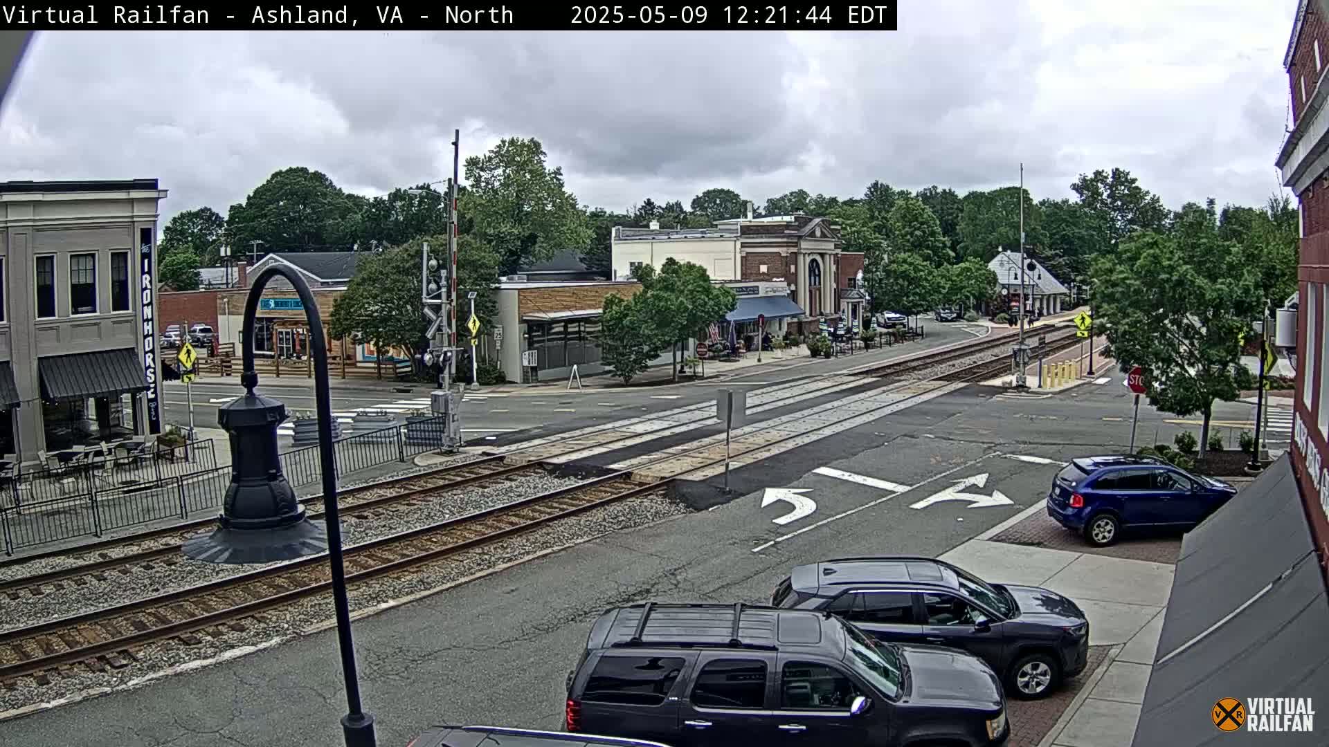 A mostly cloudy day reveals a town square with railroad tracks intersecting a street, several cars parked and driving, and buildings with awnings.