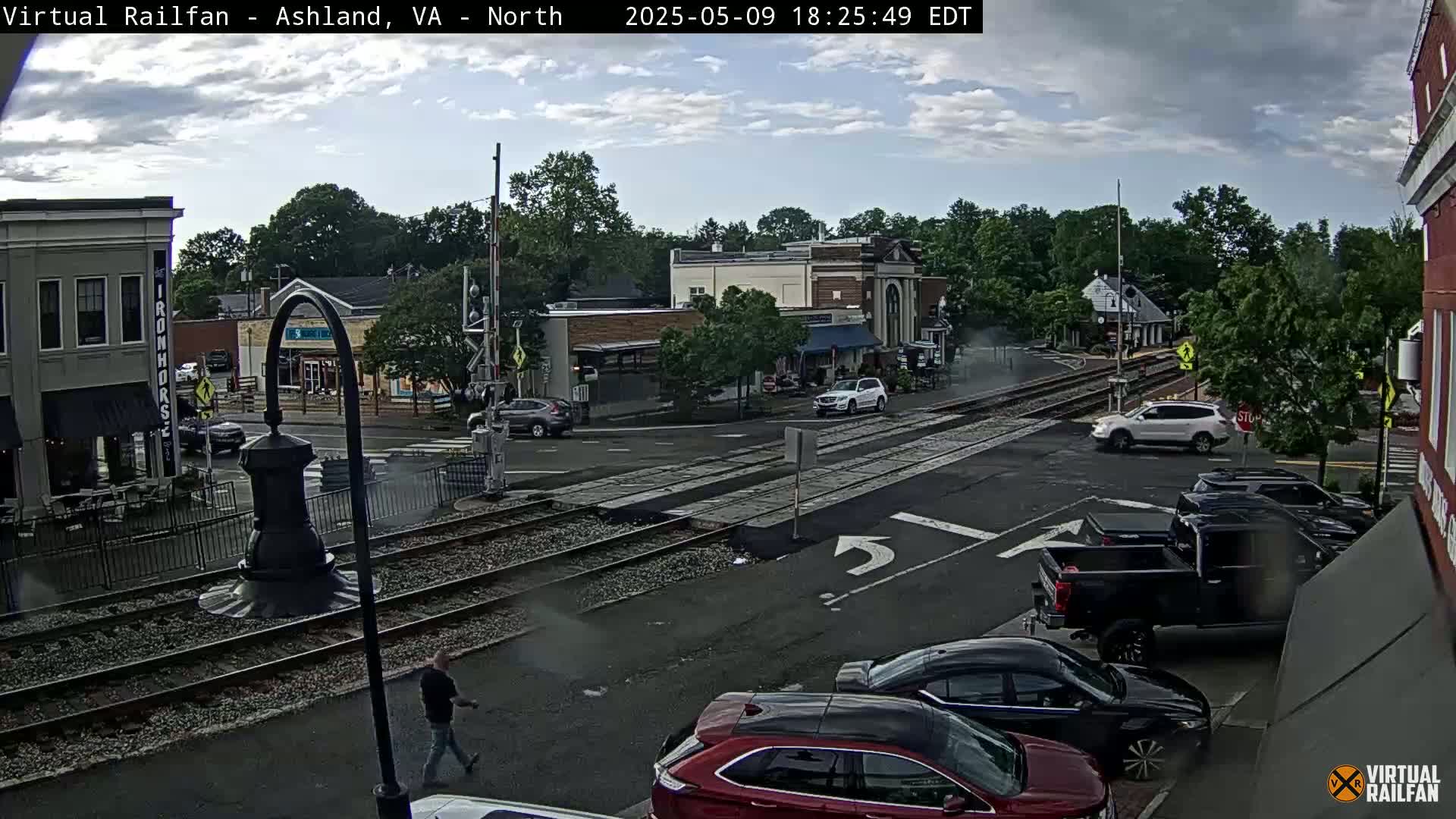 A partly cloudy day reveals a street scene with cars parked and driving near train tracks, and a pedestrian walking, all in front of a row of buildings.