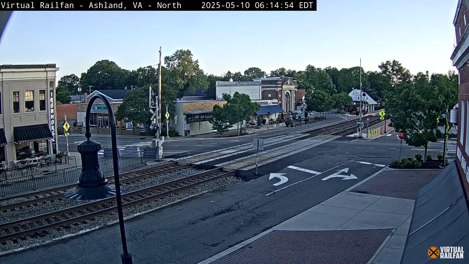 A clear morning view of a town square with railroad tracks crossing a road, featuring several buildings and lush green trees.