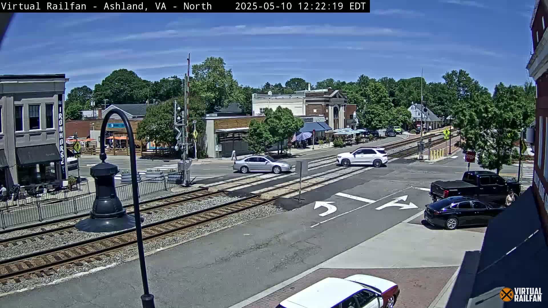 A sunny day reveals a town square with train tracks intersecting a roadway, where several cars are navigating a crossroads near various buildings and outdoor seating areas.
