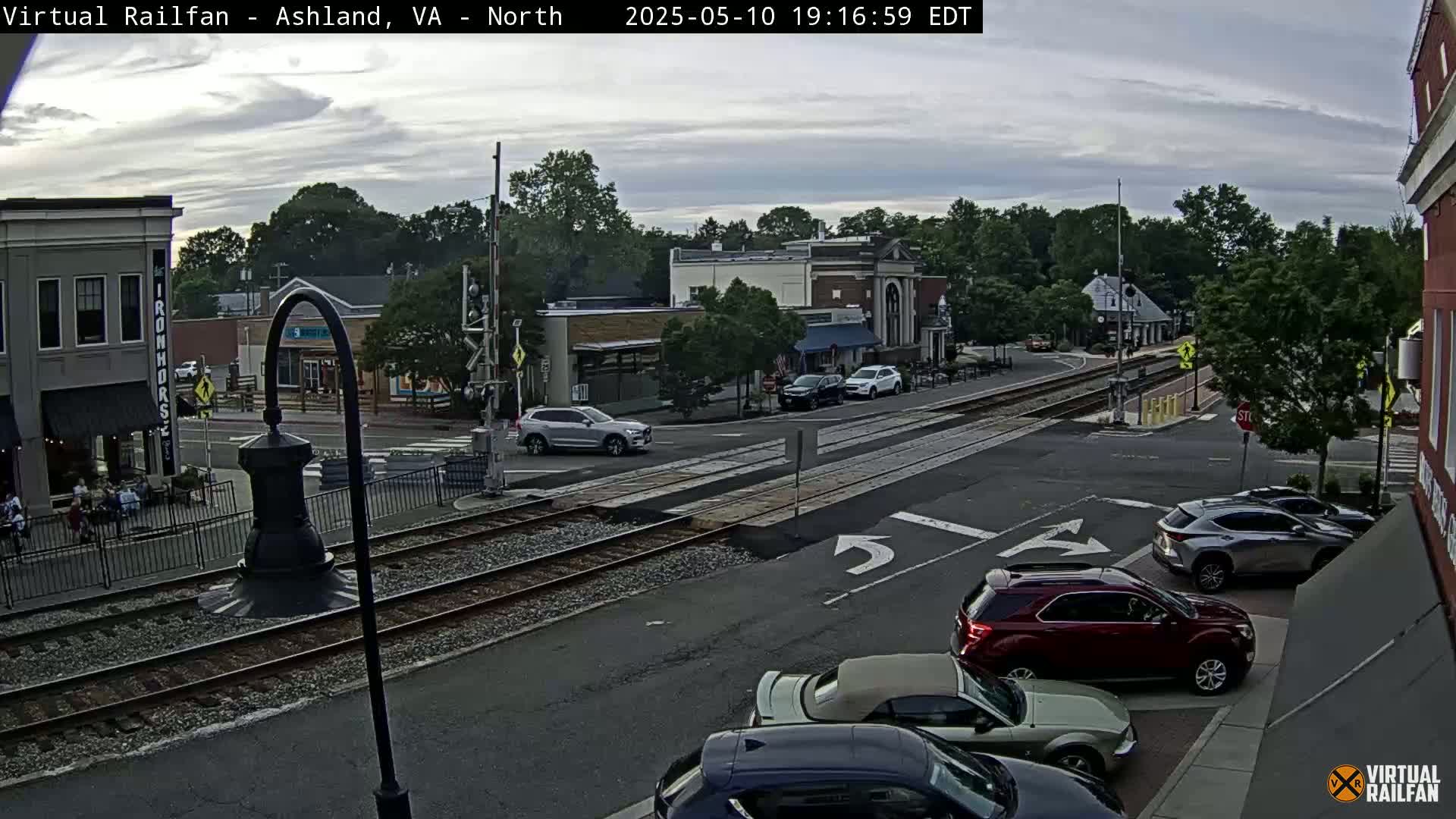 A partly cloudy evening scene shows a street intersection with several cars, train tracks, and buildings with people sitting outside.