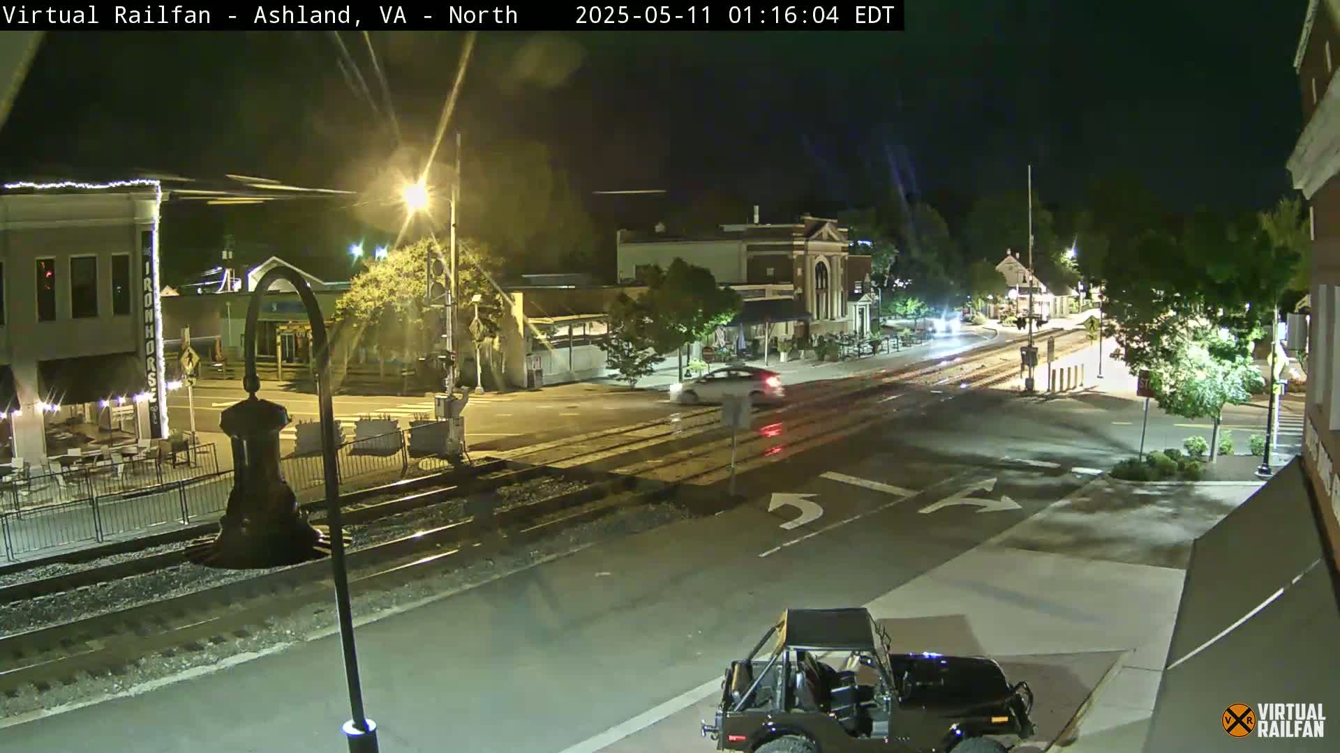 A nighttime view of a town square with a car driving through the intersection, train tracks visible in the foreground, and streetlights illuminating the scene.