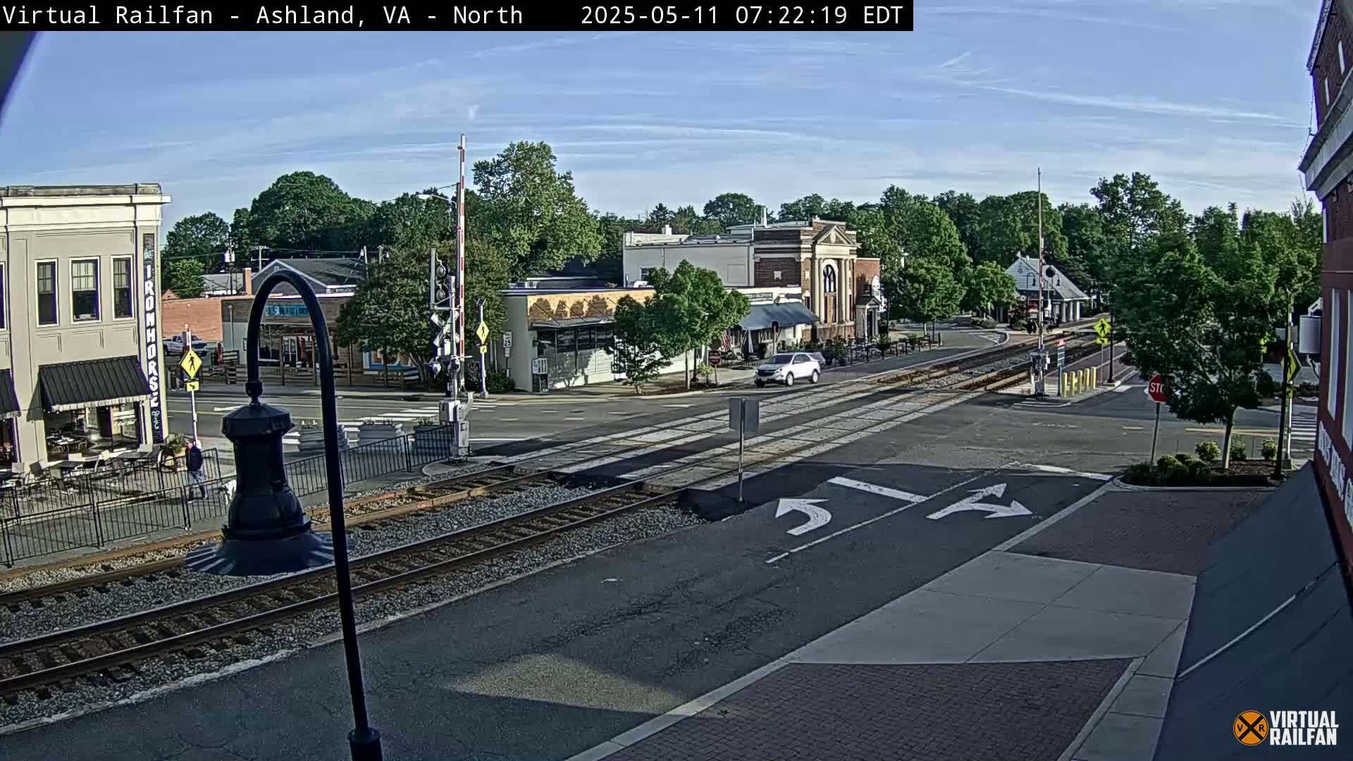 A sunny day in a small town features a railroad crossing in the center of the intersection, with several buildings and some cars visible.