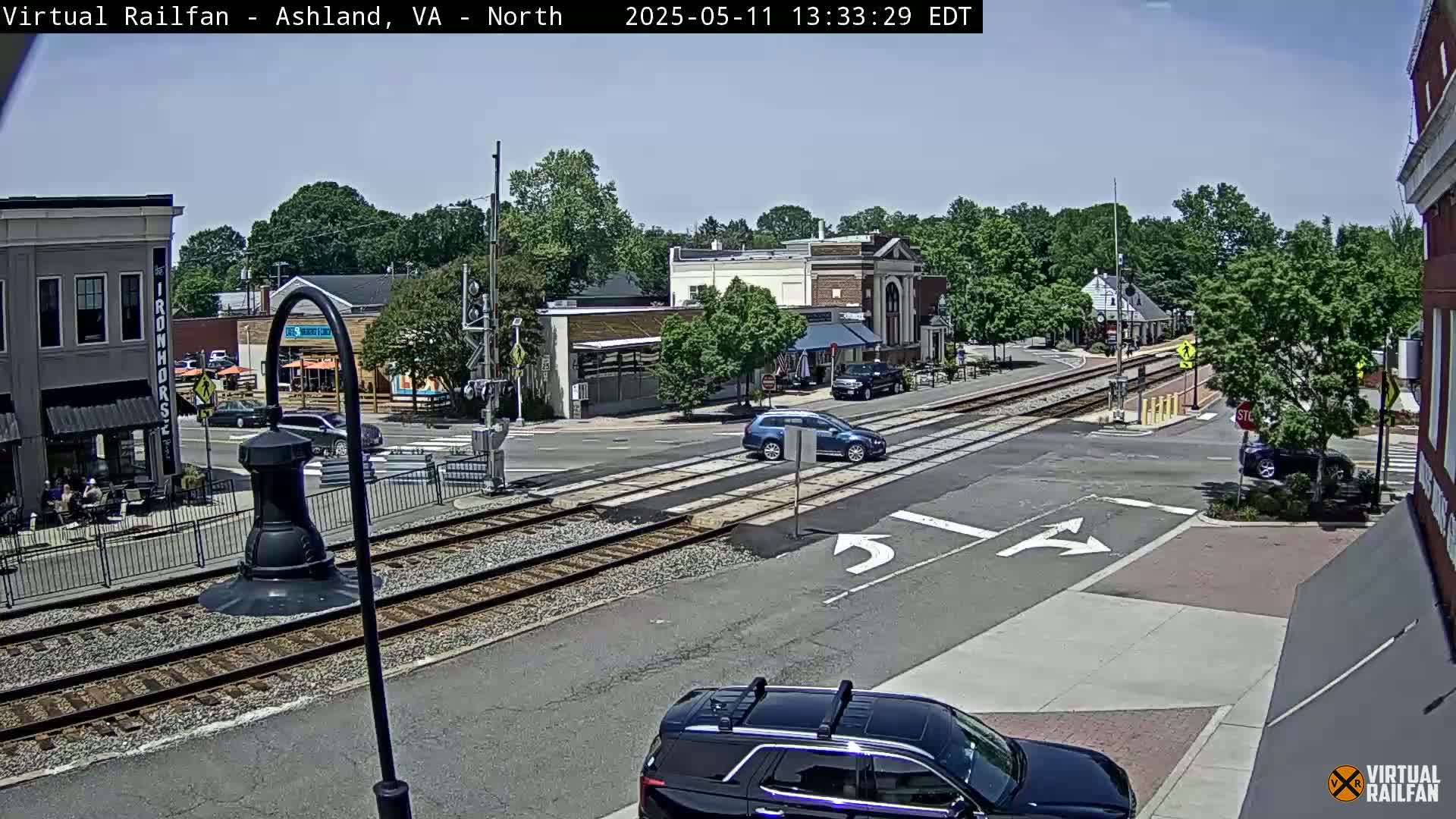A sunny day view of a town intersection with railroad tracks, showing several cars and buildings, with some people sitting outdoors at a cafe.