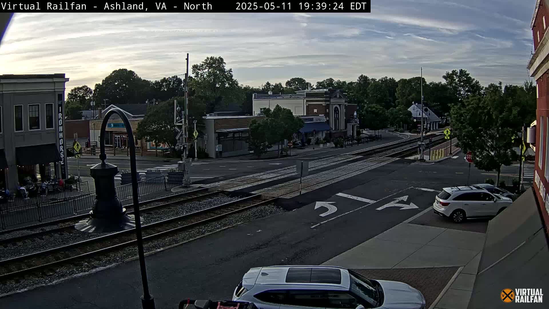 A mostly cloudy evening scene shows a street intersection with railroad tracks, several buildings, and a few cars, one of which is silver.