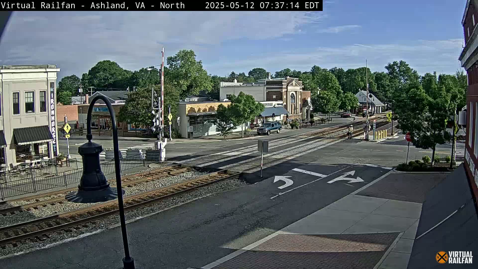A mostly sunny view of a town's street intersection with railroad tracks, featuring several buildings and a vehicle, under a partly cloudy sky.