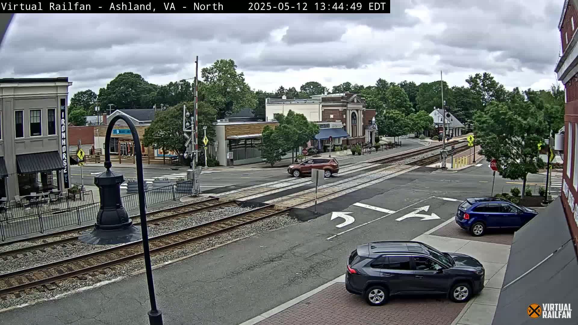 A partly cloudy day shows a railroad crossing in a town with several cars at an intersection near brick buildings and trees.