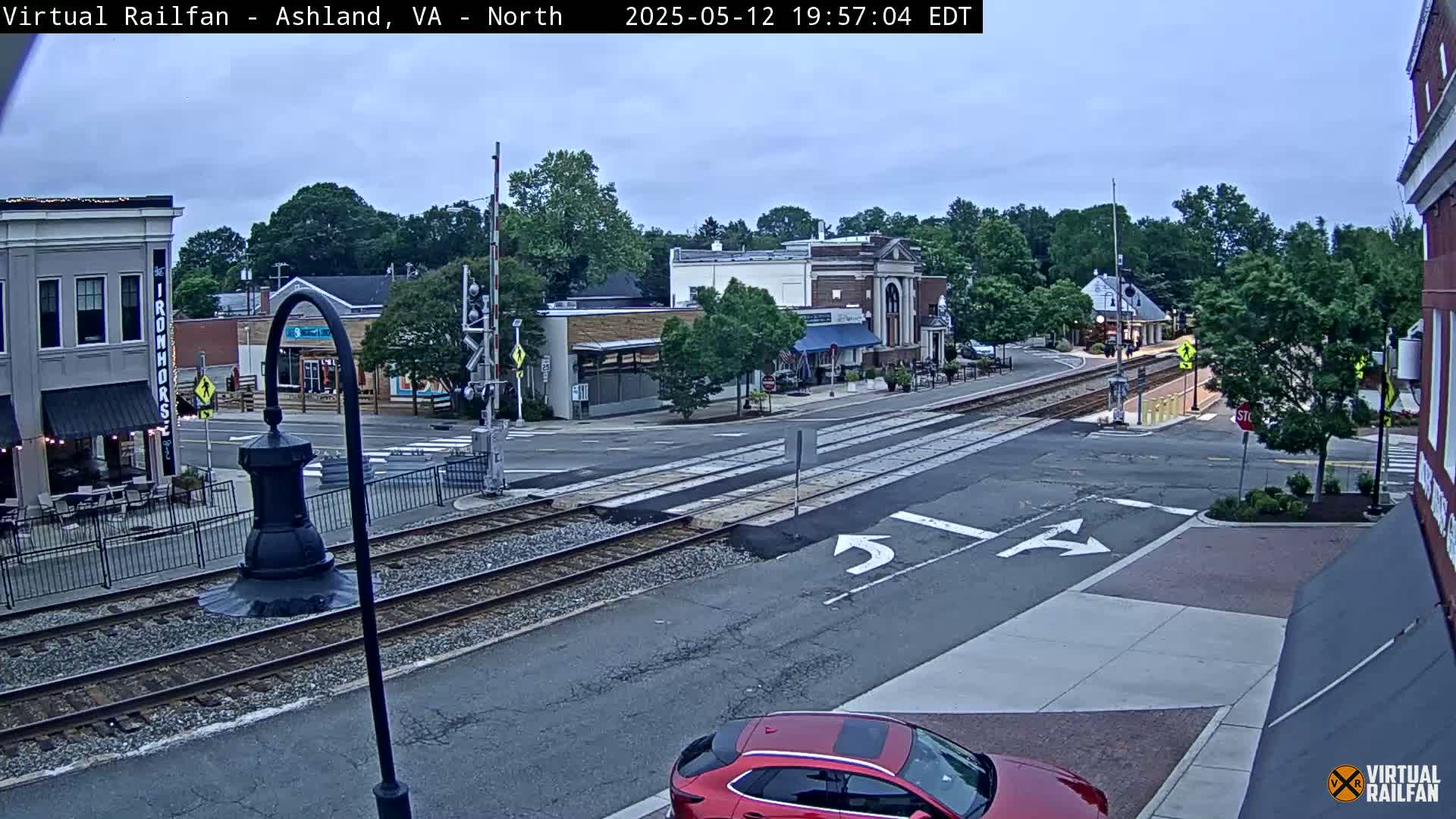 A mostly cloudy daytime scene shows a street intersection with railroad tracks crossing, several buildings, and a red car.