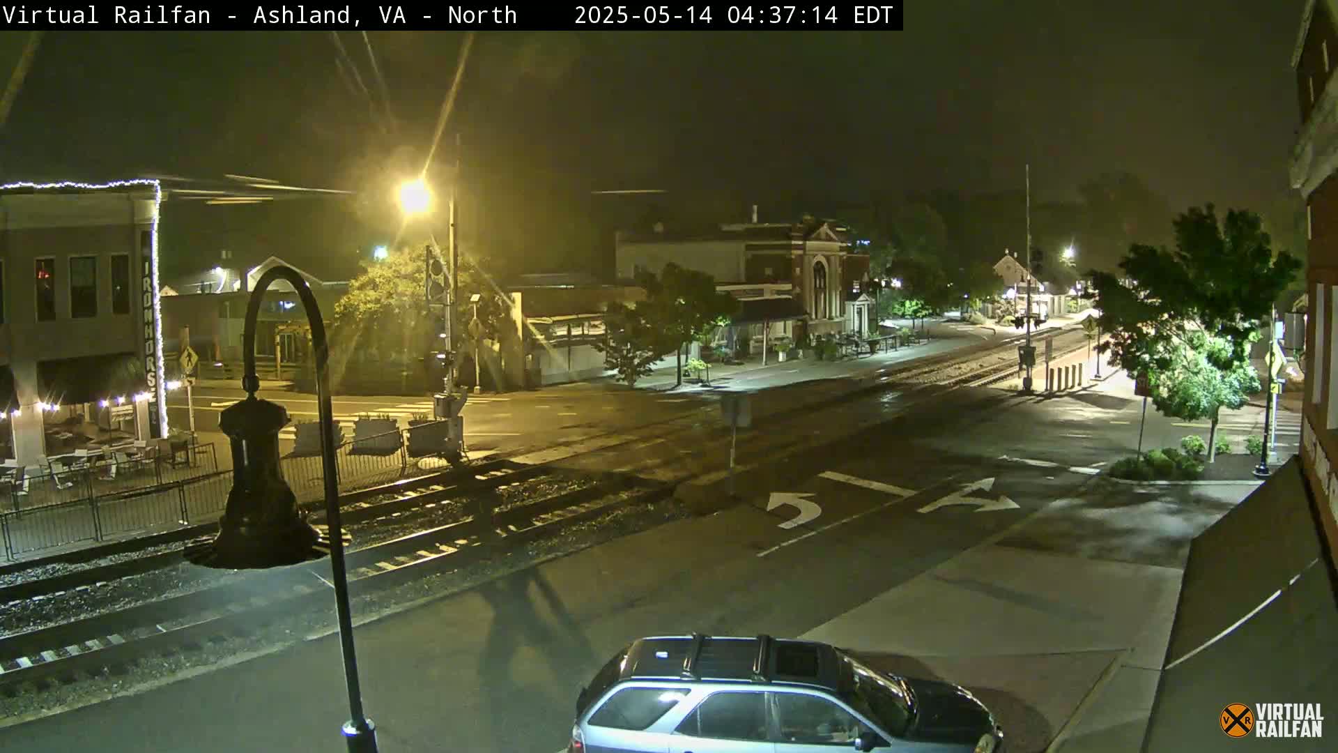 A nighttime street scene shows a wet road intersecting with train tracks, passing in front of buildings and a parked SUV under dim streetlights.