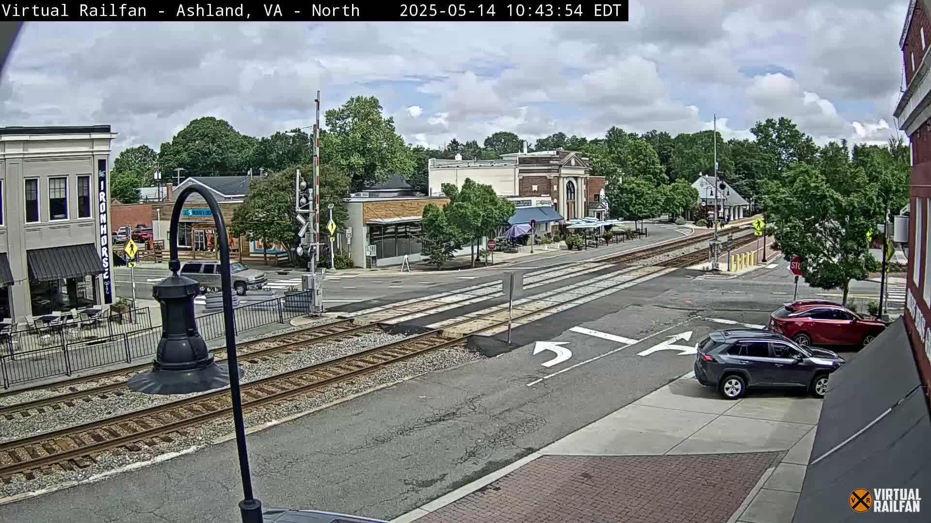 A partly cloudy day reveals a town square with railroad tracks crossing a street, several cars parked and driving, and buildings with outdoor seating.