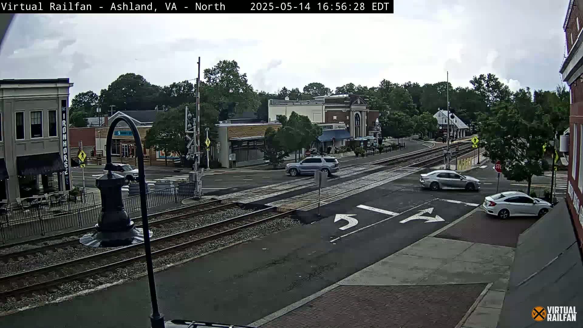 A mostly cloudy day shows a street intersection with several cars, railroad tracks, and several buildings in a town.