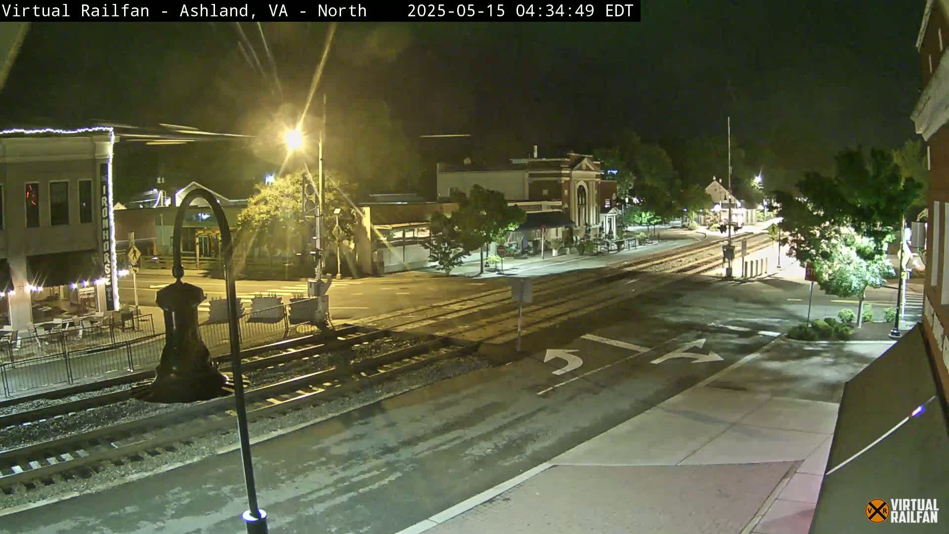 A nighttime street scene shows a railroad crossing intersecting a road in a town with buildings lit along the street under a clear sky.