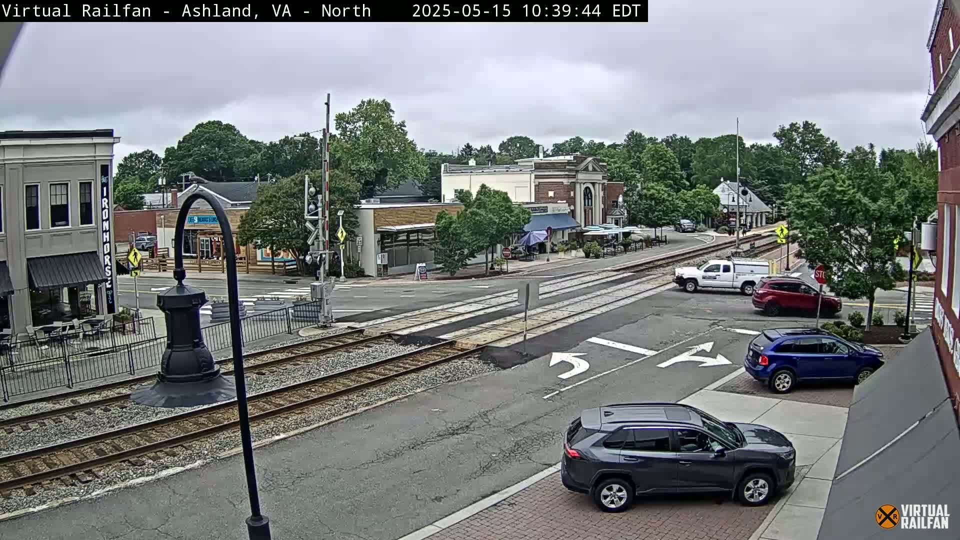 A mostly cloudy day shows a street scene with several cars parked and driving near train tracks that cross a town square with buildings and outdoor seating.