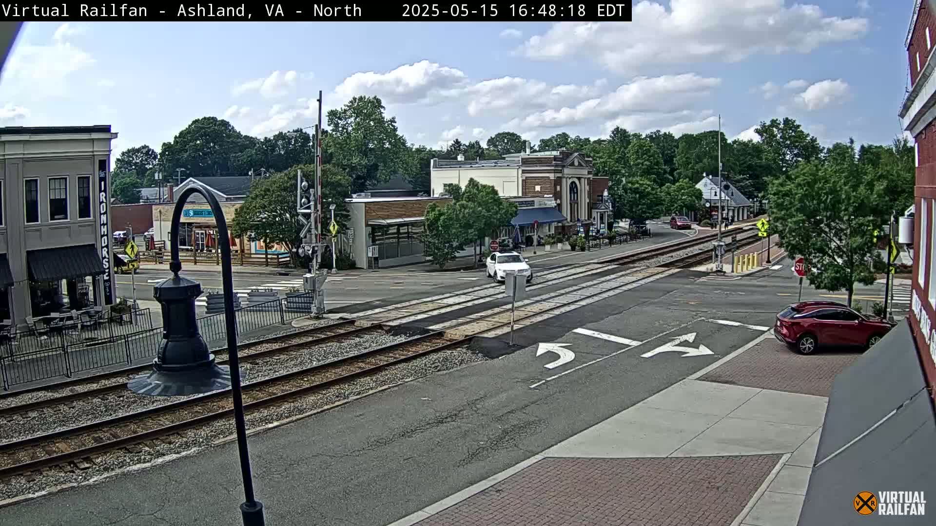 A partly cloudy day reveals a street intersection with railroad tracks, several buildings, and a few cars, one of which is red.