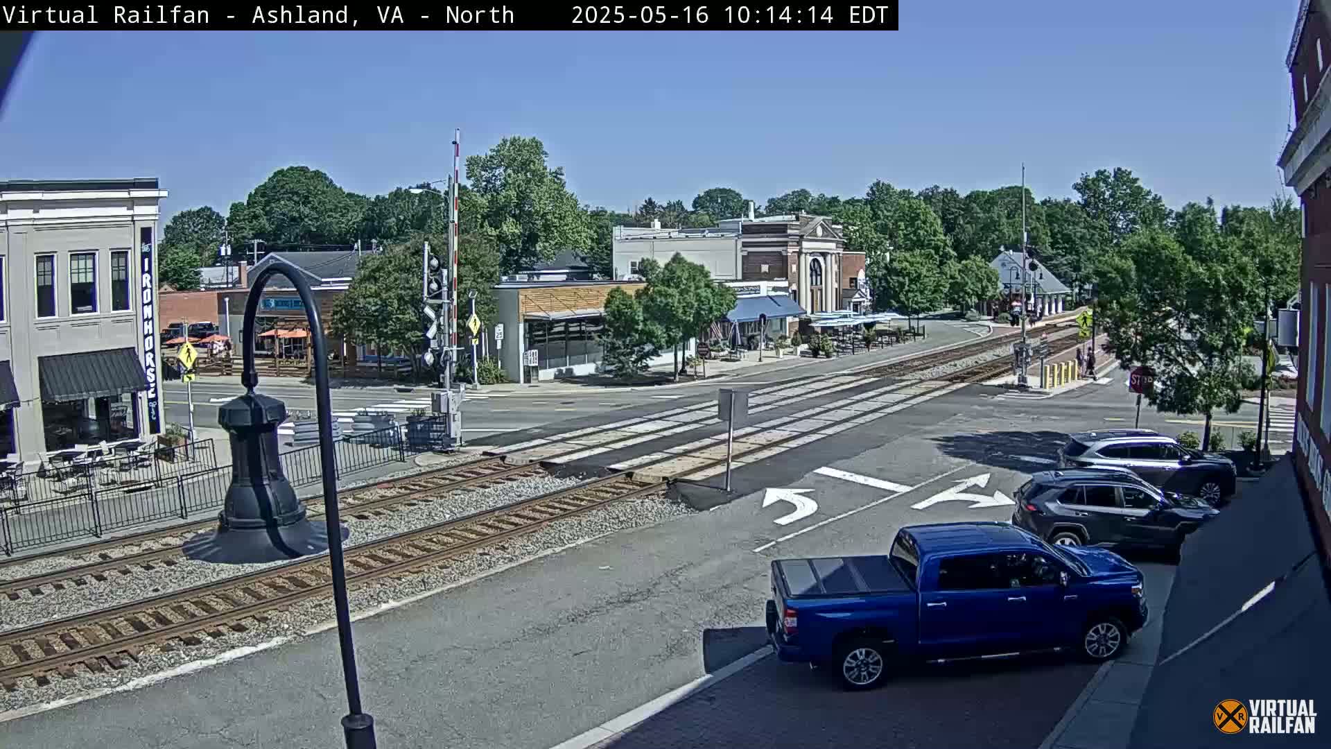 A sunny day shows a railroad crossing in a town with several buildings, vehicles, and outdoor seating areas.