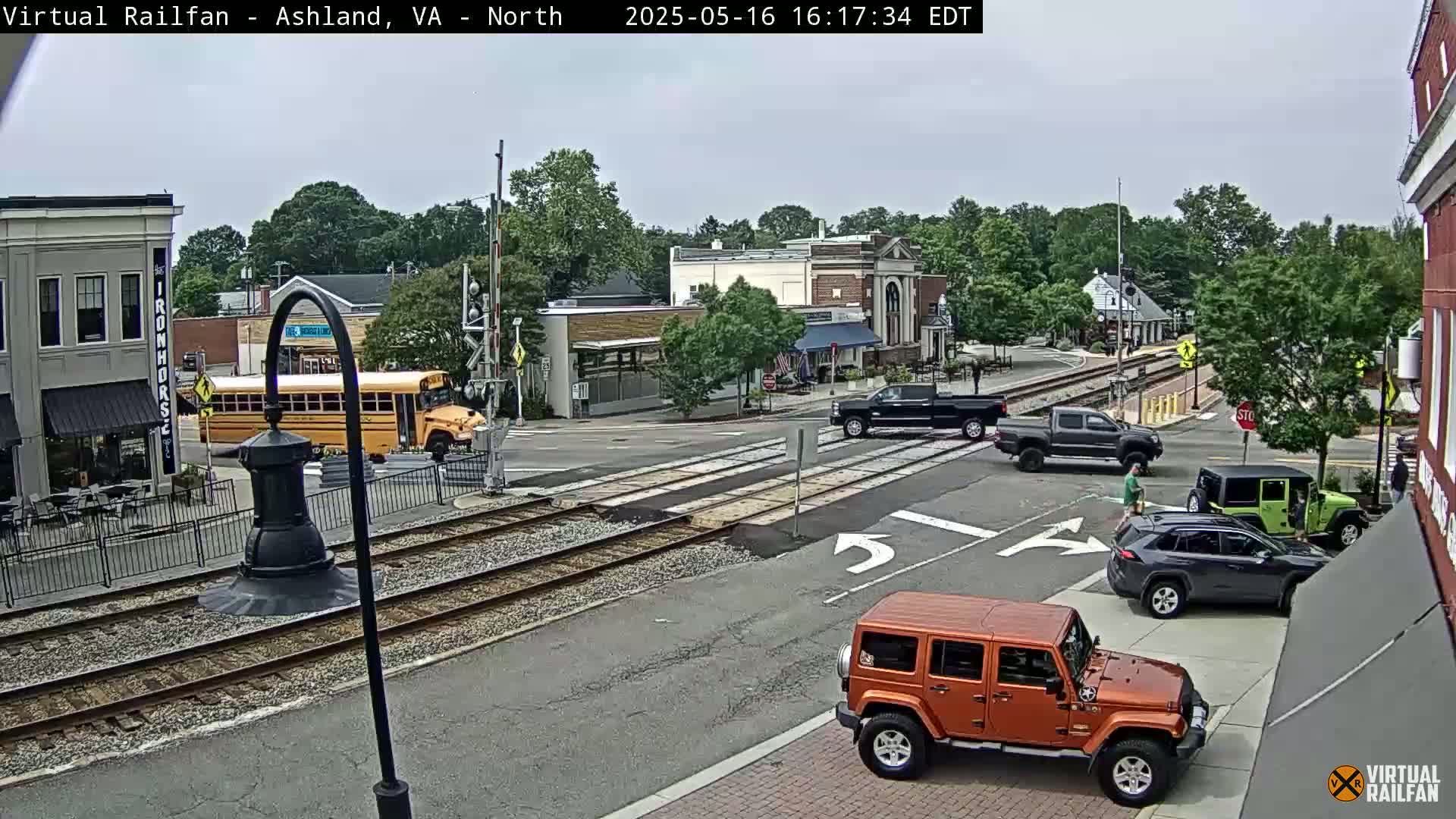 A partly cloudy day shows a town square with several vehicles at an intersection crossing train tracks, including a school bus, pickup truck, Jeep, and SUV.